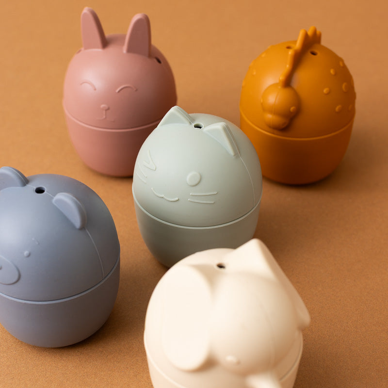 Five small egg-shaped cat-themed containers in pastel colors, including pink, orange, blue-gray, mint green, and cream, arranged on a warm brown background.