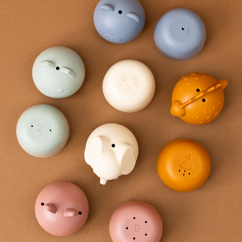 A set of cute, round silicone charging cases arranged on a textured brown background. The cases come in various pastel and bright colors, including light blue, dusty blue, mint green, cream, orange, and pink, each with a small animal or character face etched on the front.