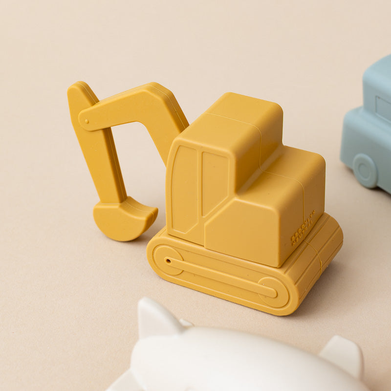 A yellow toy excavator with a scoop arm, photographed on a warm beige surface. Part of a light blue toy vehicle is visible on the right, and the top of a white toy is visible at the bottom.