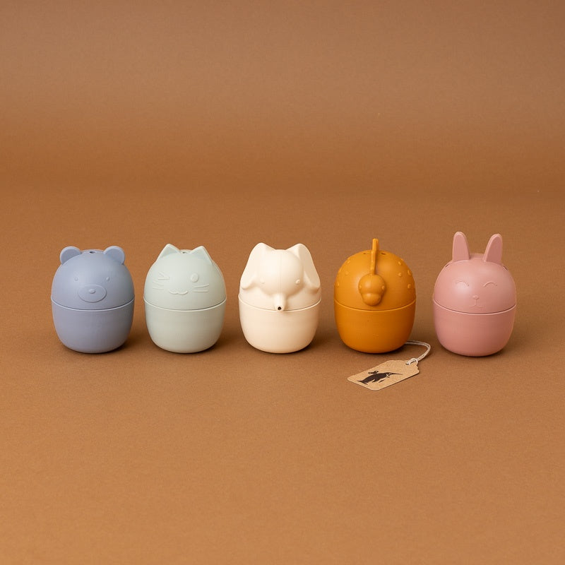 Five small rounded capsule-shaped containers in pastel and earthy colors, arranged in a row on a warm brown background. The designs resemble animal or character heads, including bear, cat, fox, duck, and bunny motifs.