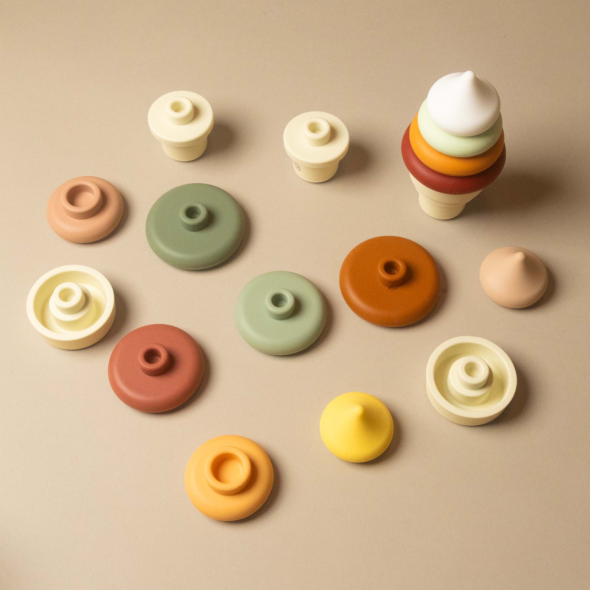A set of pastel-colored silicone-ice-cream-cone-stacker-set baby teething toys and lids in various shapes, including a cone-shaped ice cream toy, arranged on a neutral beige background.