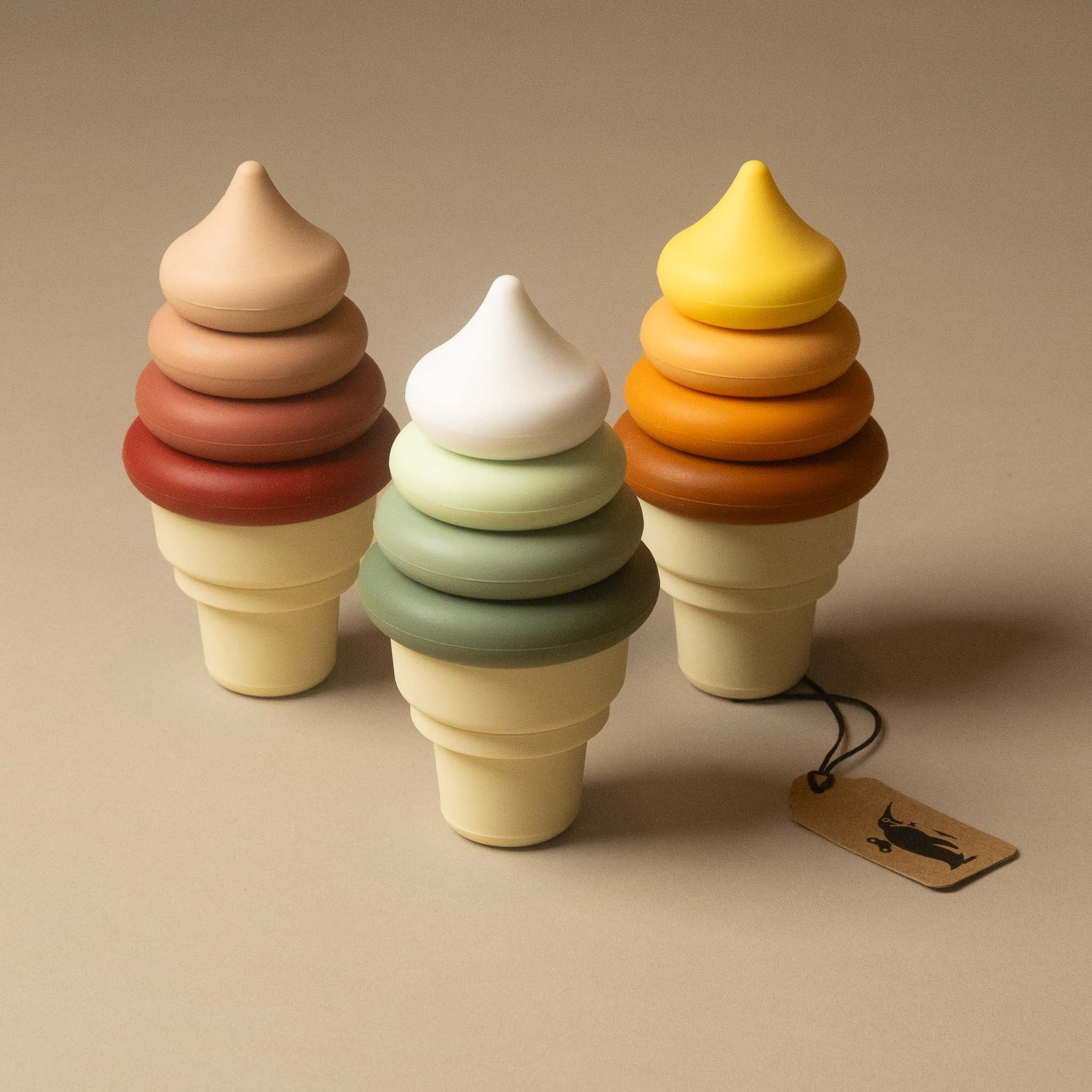 Three silicone-ice-cream-cone-stacker-in set in stacked pastel and warm tones, arranged on a beige studio background. A small hanging tag is attached to the rightmost cone.