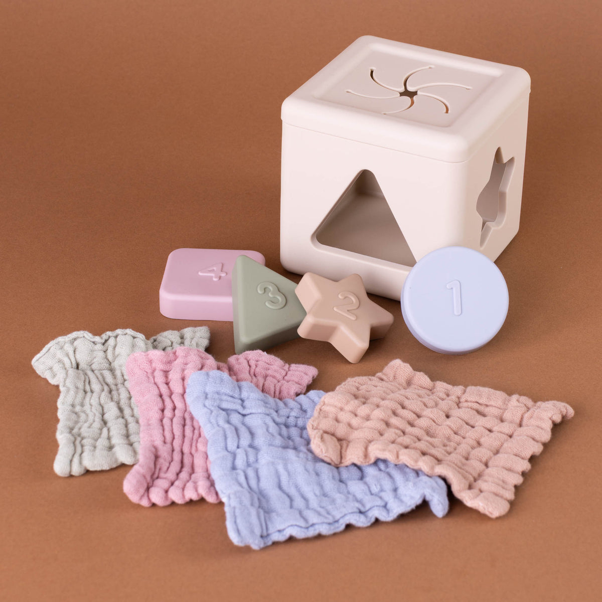 A baby sensory sorting toy set on a warm brown background, featuring a cream-colored sorting box, pastel-colored shape and number blocks, and soft textured cloth swatches in light gray, pink, blue, and peach.