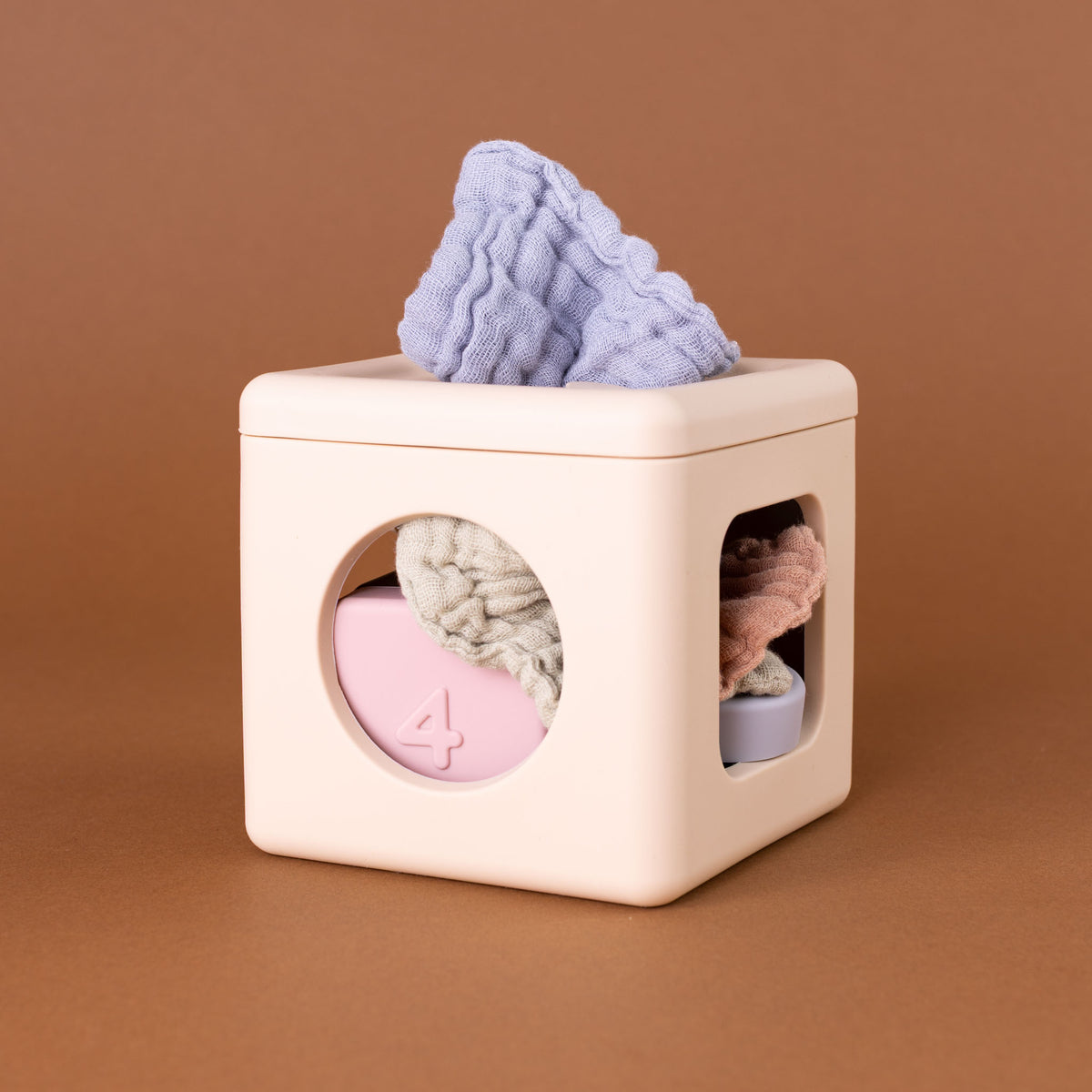 A cream-colored cube-shaped baby tissue box with circular and square cutouts, holding soft pastel baby tissues in pink, lavender, and beige, set against a solid brown background.