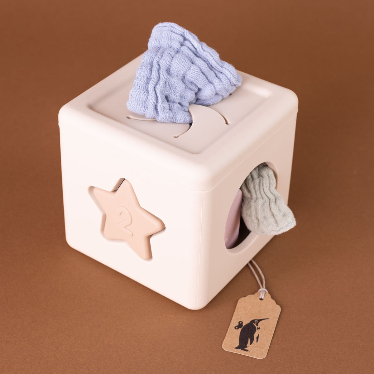 A cream-colored wooden toy box with a star-shaped slot and a circular cutout, shown on a brown background. A light blue cloth is sticking out of the top slot, and a pale green cloth is visible in the side cutout.