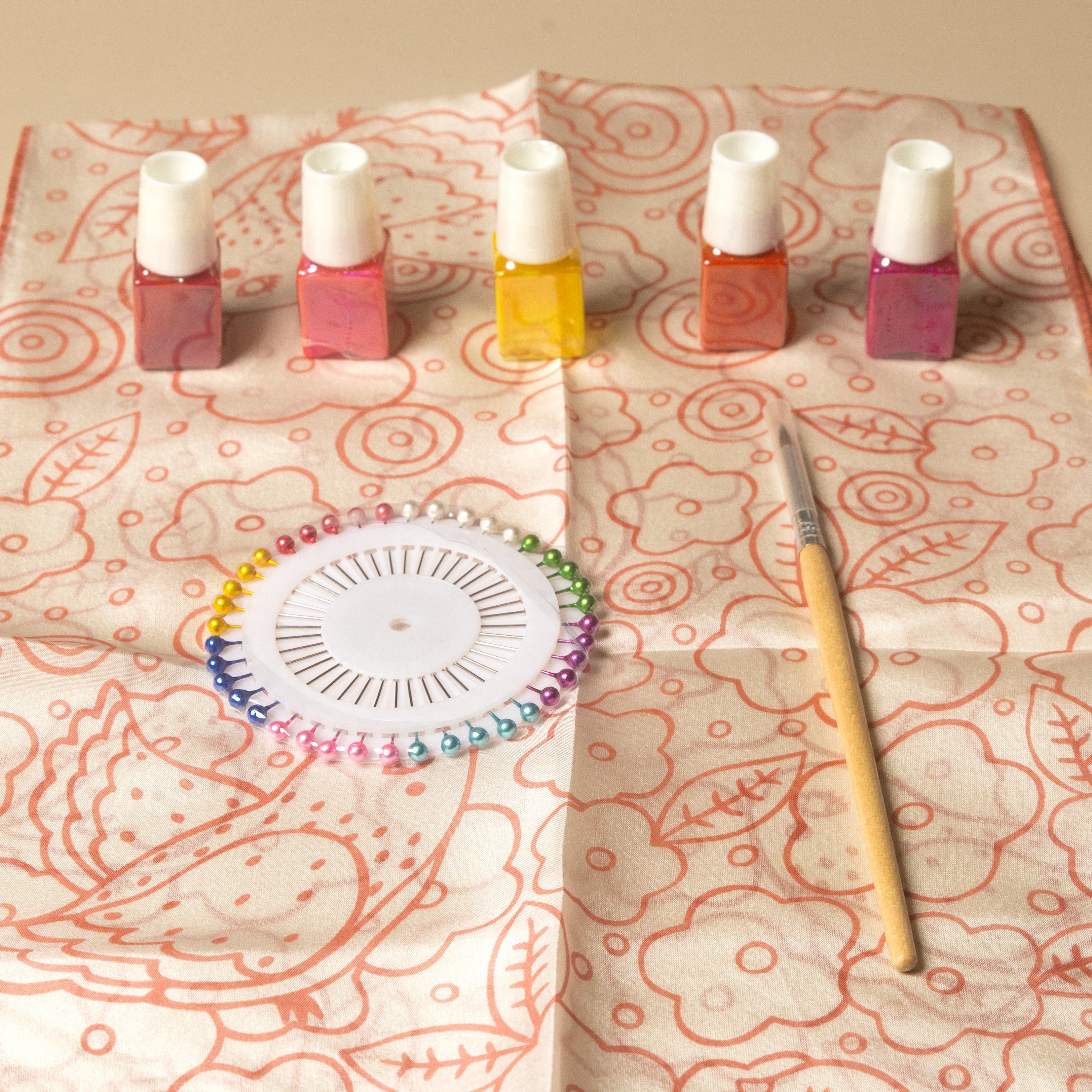 A flat-lay of craft supplies on patterned paper, including five small bottles of nail polish in pink, orange, and yellow shades, a circular bead organizer, and a wooden-handled paintbrush.