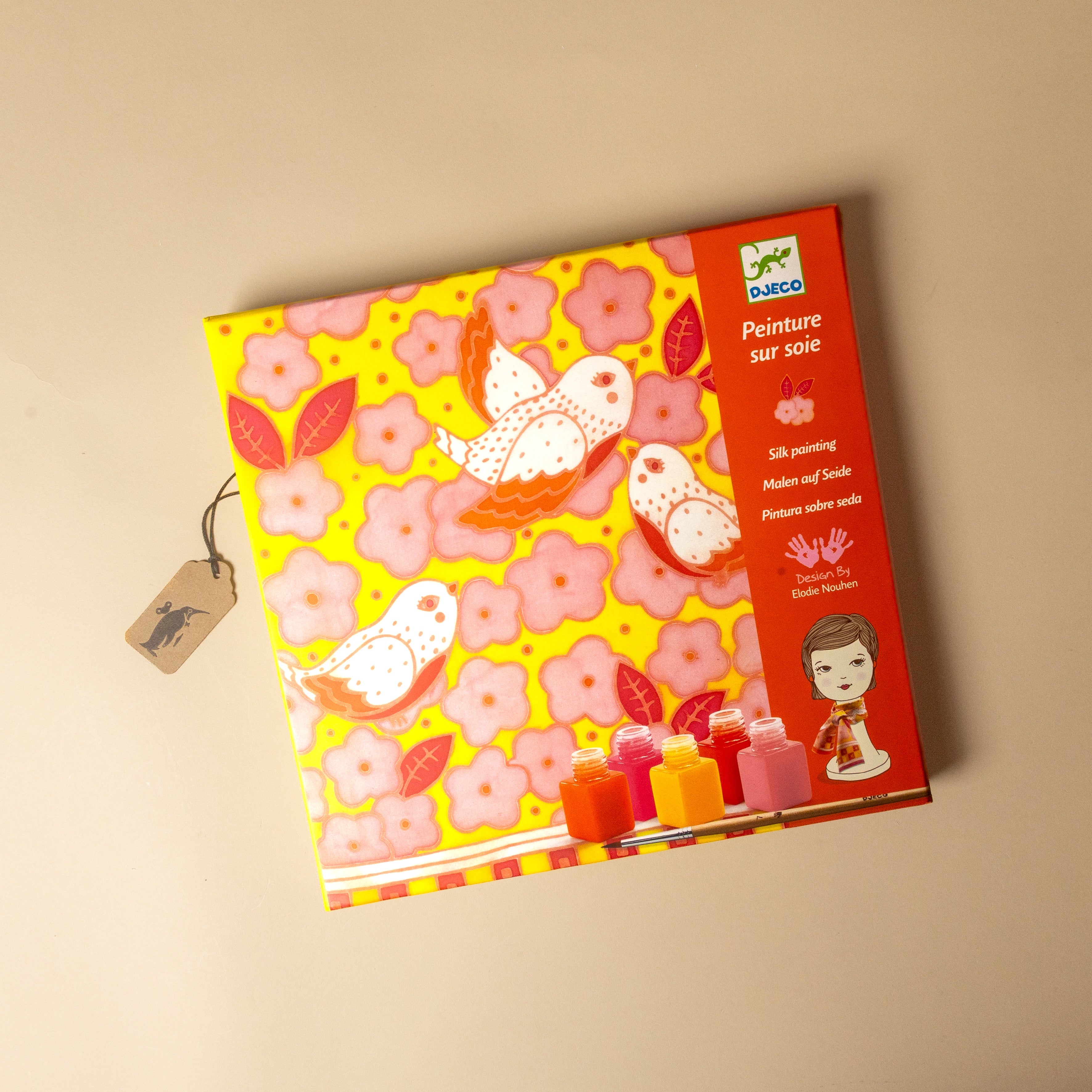 A boxed children's painting , titled 'Peinture sur soie', shown on a plain beige background. The box features a bright yellow and pink floral pattern with white birds, a red side panel with product details and a child illustration, and a small hanging tag.