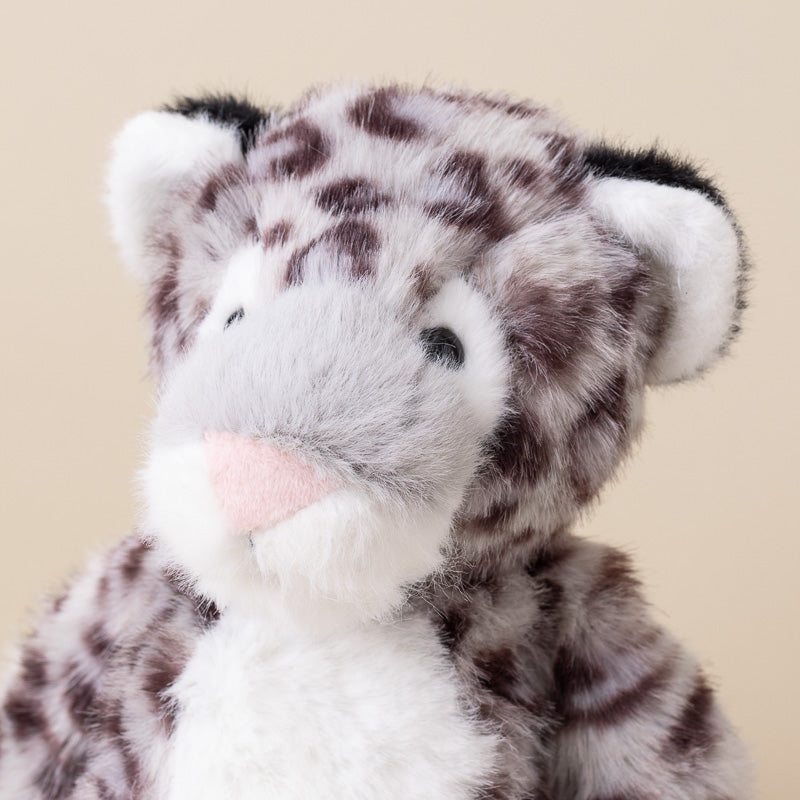 A close-up of a plush stuffed animal with a spotted feline pattern, featuring gray and white fur, dark brown spots, a pink nose, and white inner ears, set against a plain beige background.