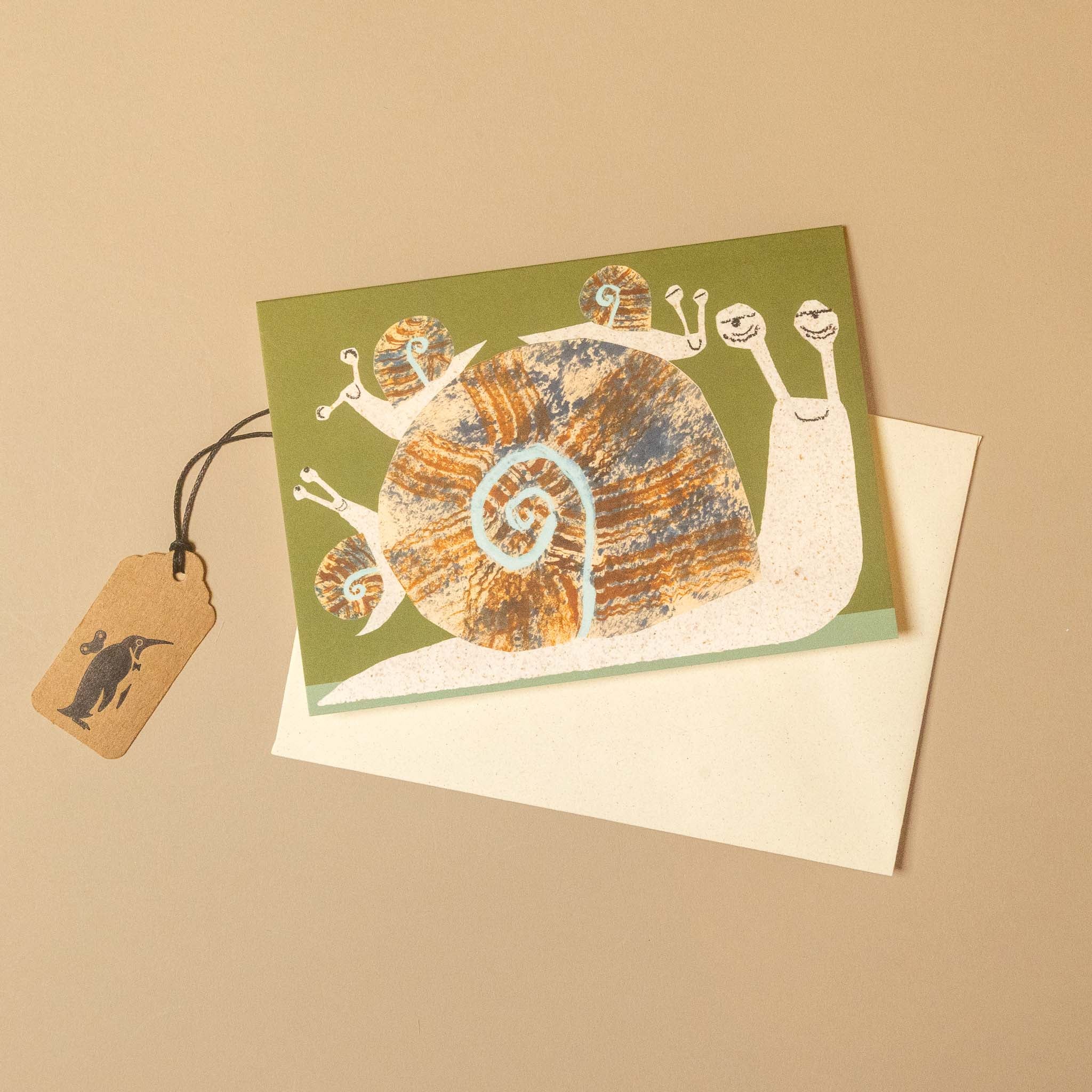 A square greeting card with a whimsical snail illustration on an olive green background, placed on top of a white envelope. A small kraft paper hangtag with a black snail silhouette is attached by a black string.