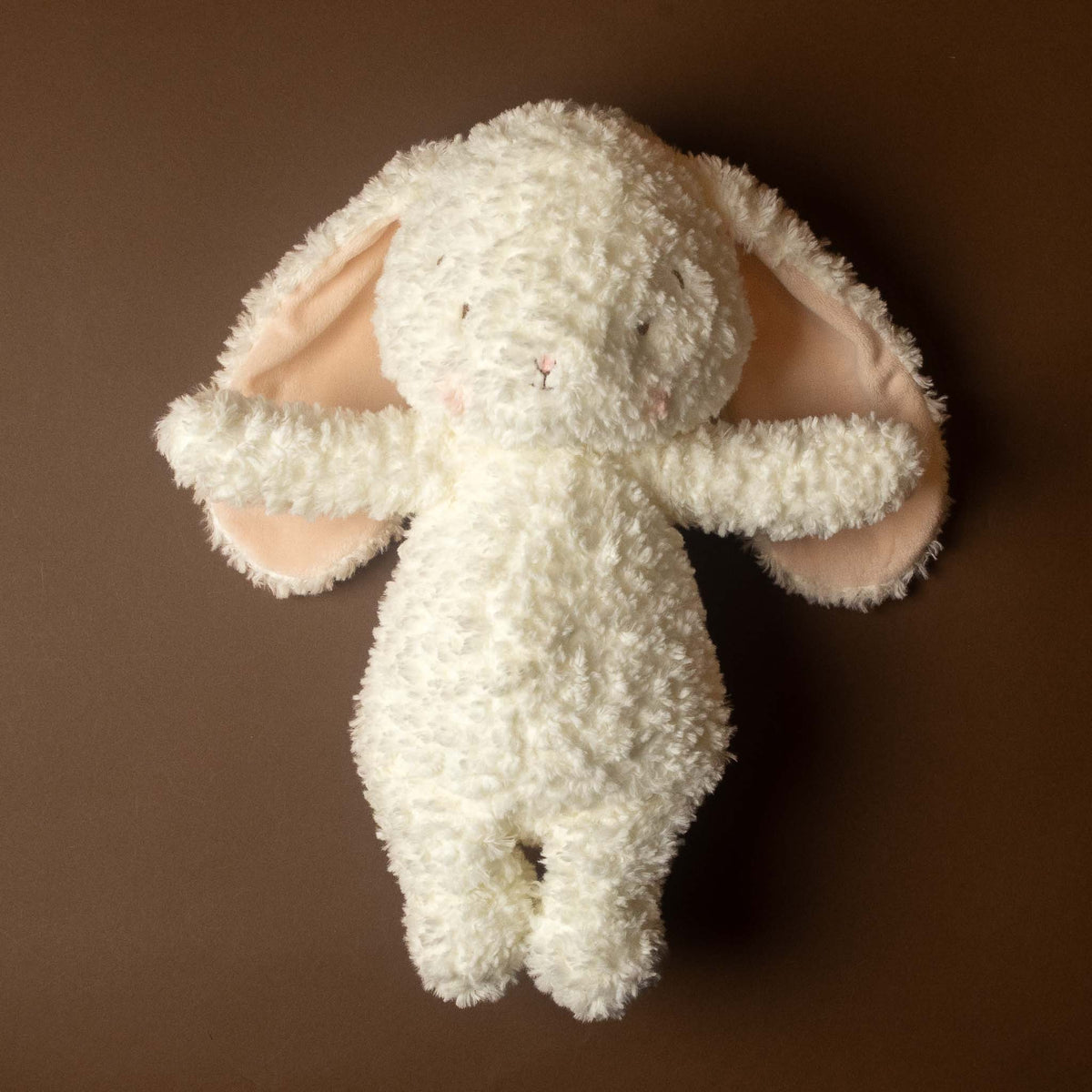 A fluffy snowball-the-white-bunny-stuffed-animal with long floppy ears and pale pink inner ears, photographed against a solid dark brown background.