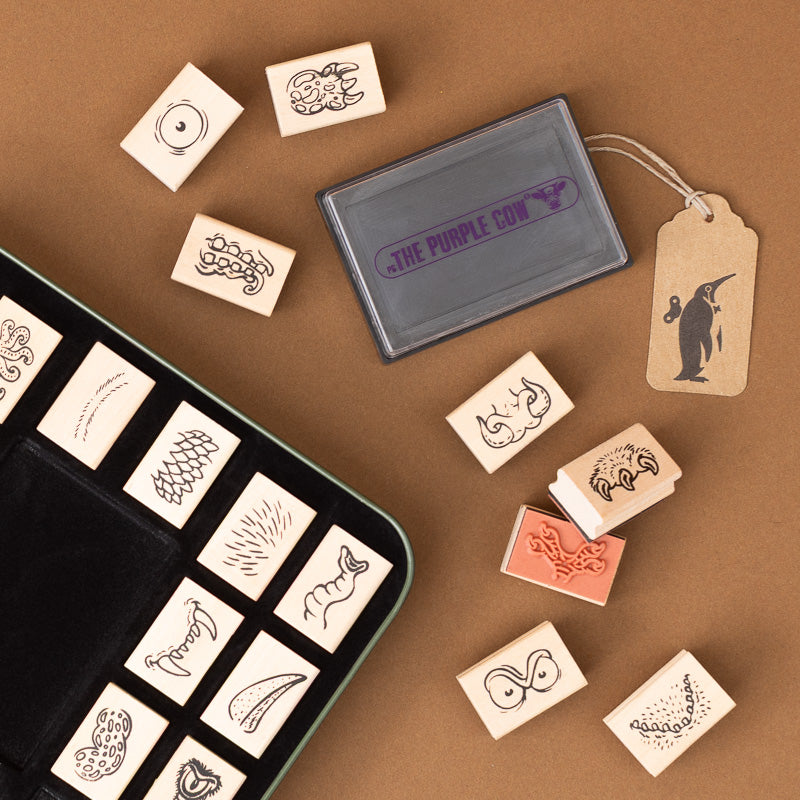 A flat-lay of rubber stamp set accessories on a brown background, including a gray ink pad several wooden stamps with black animal and nature motifs, a kraft paper hang tag, and a black case with additional stamps.