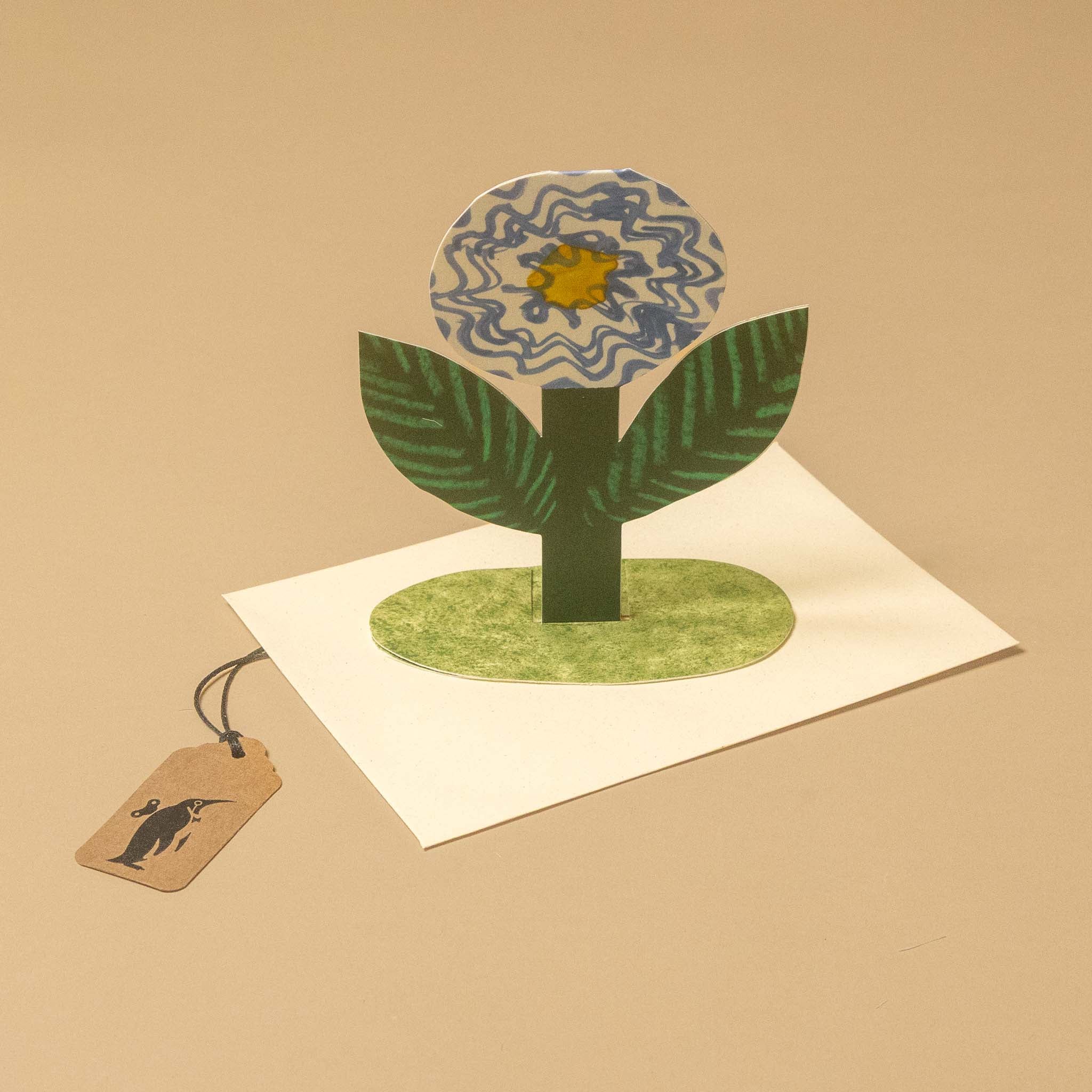 A handmade 3D pop-up flower card on a beige background. The card features a green stem and leaves with a blue and yellow patterned flower head, set on a green textured circular base over an open white card.
