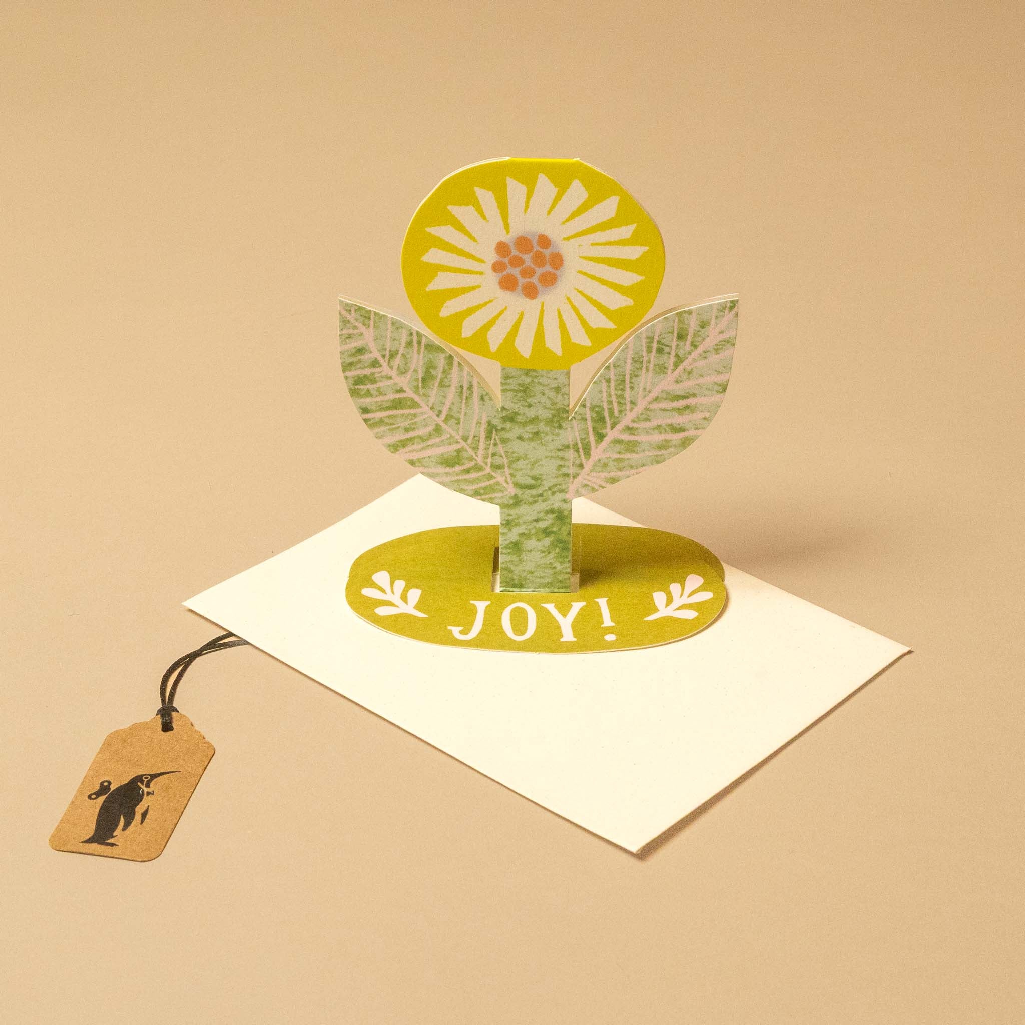 A 3D pop-up paper flower greeting card on a white envelope, featuring a yellow and white flower with green leaves on a beige background. A small paper hangtag with a black bird silhouette is attached to the left side.