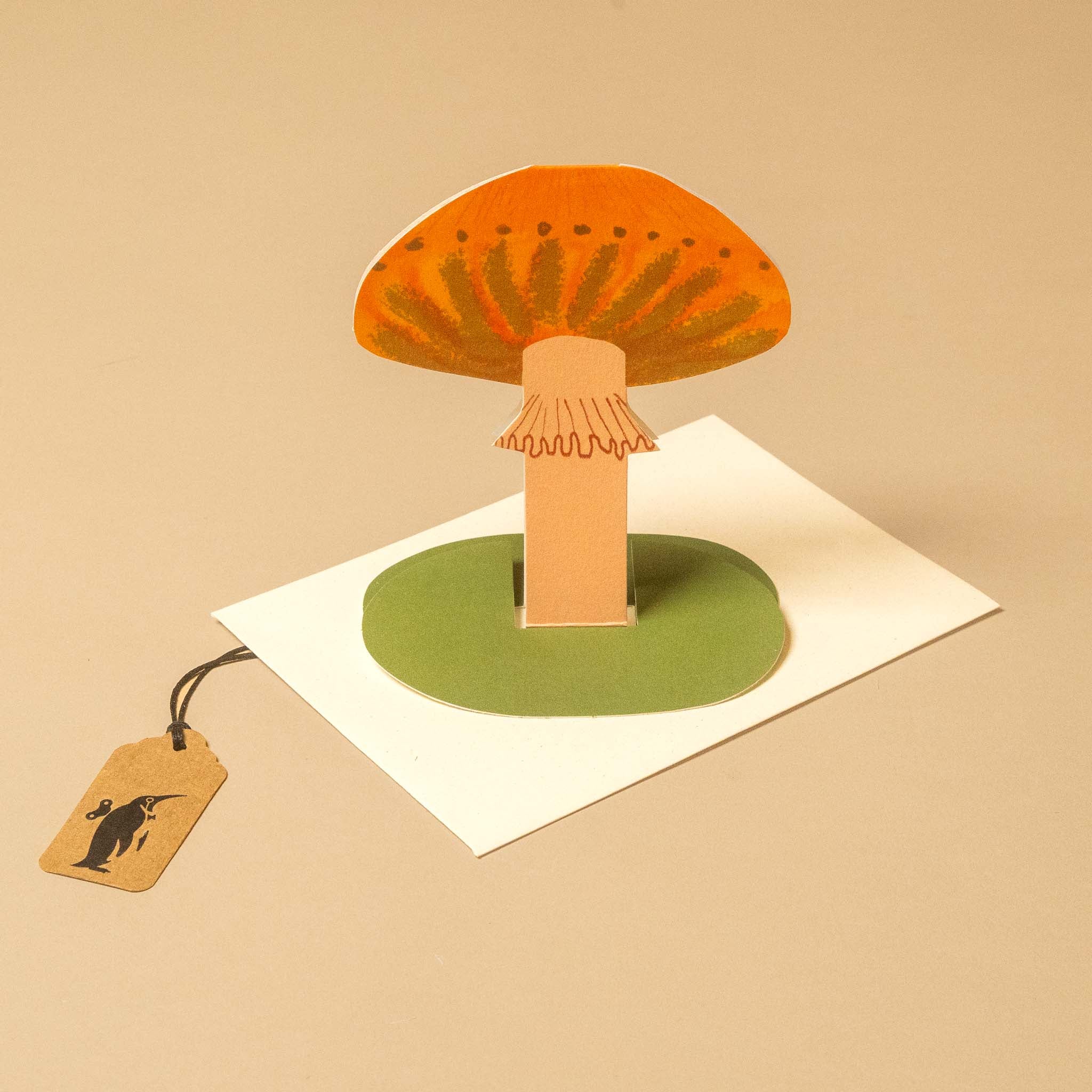 A 3D pop-up mushroom card featuring an orange cap with green spots, a tan stem, and a green circular base, displayed on a white square card against a beige background with a small hanging tag.