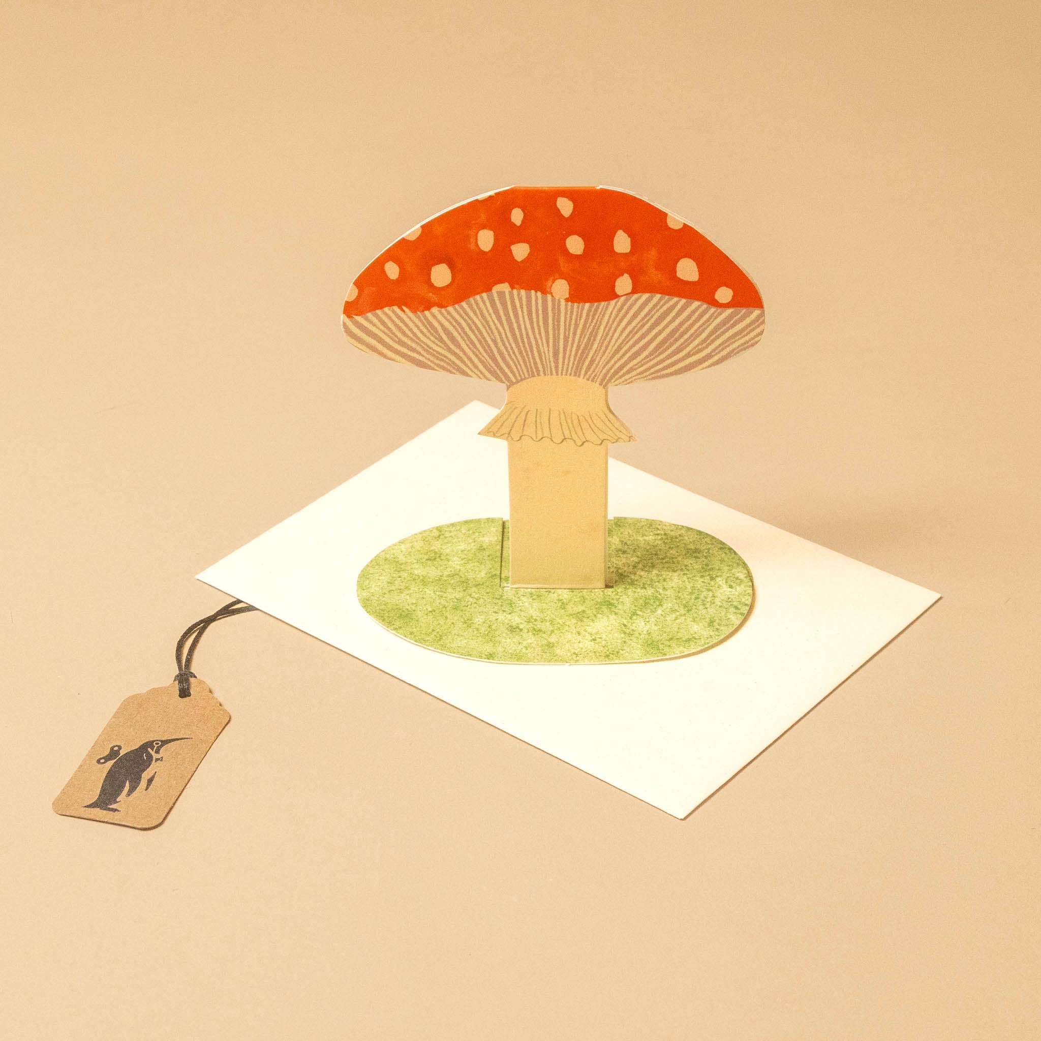 A 3D pop-up mushroom card featuring a red and white spotted cap on a green circular base, mounted on a white square card with a hanging paper tag, set against a solid beige background.