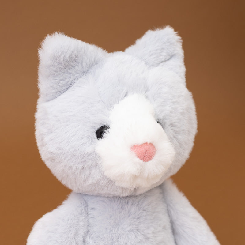 A close-up of a plush toy shaped like a cat, with light gray fur, a white muzzle, a small pink nose, and black embroidered eyes, set against a solid warm brown background.
