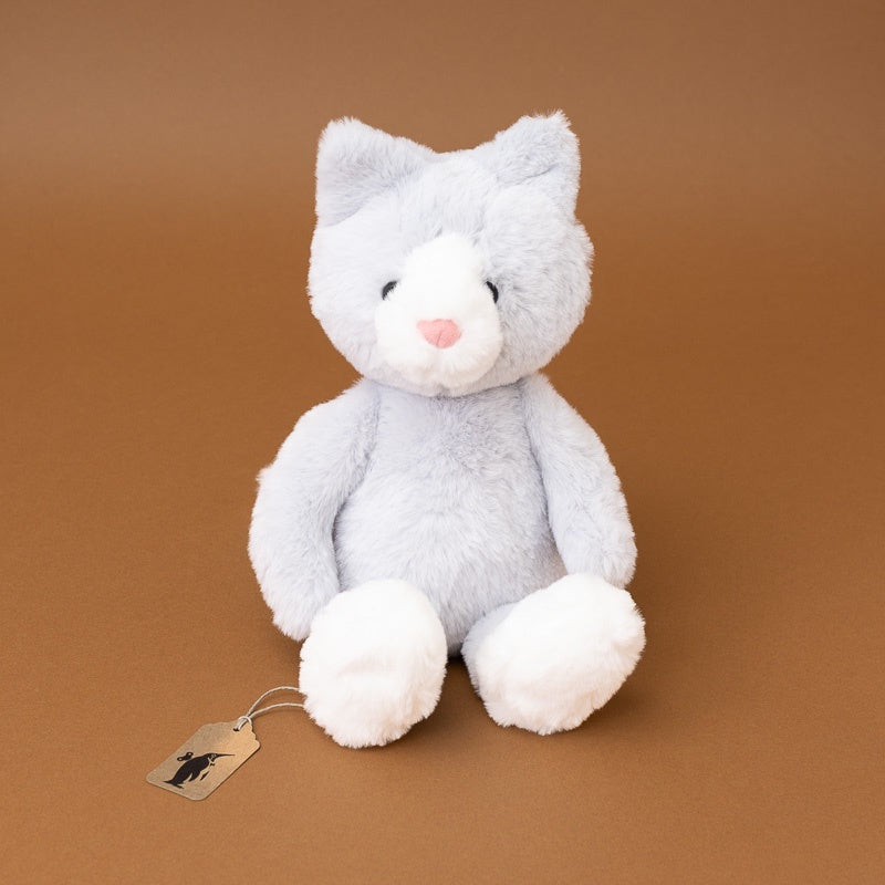 A light gray and white plush cat toy with a small pink nose, sitting on a smooth brown background. A small paper tag is attached near the left side.