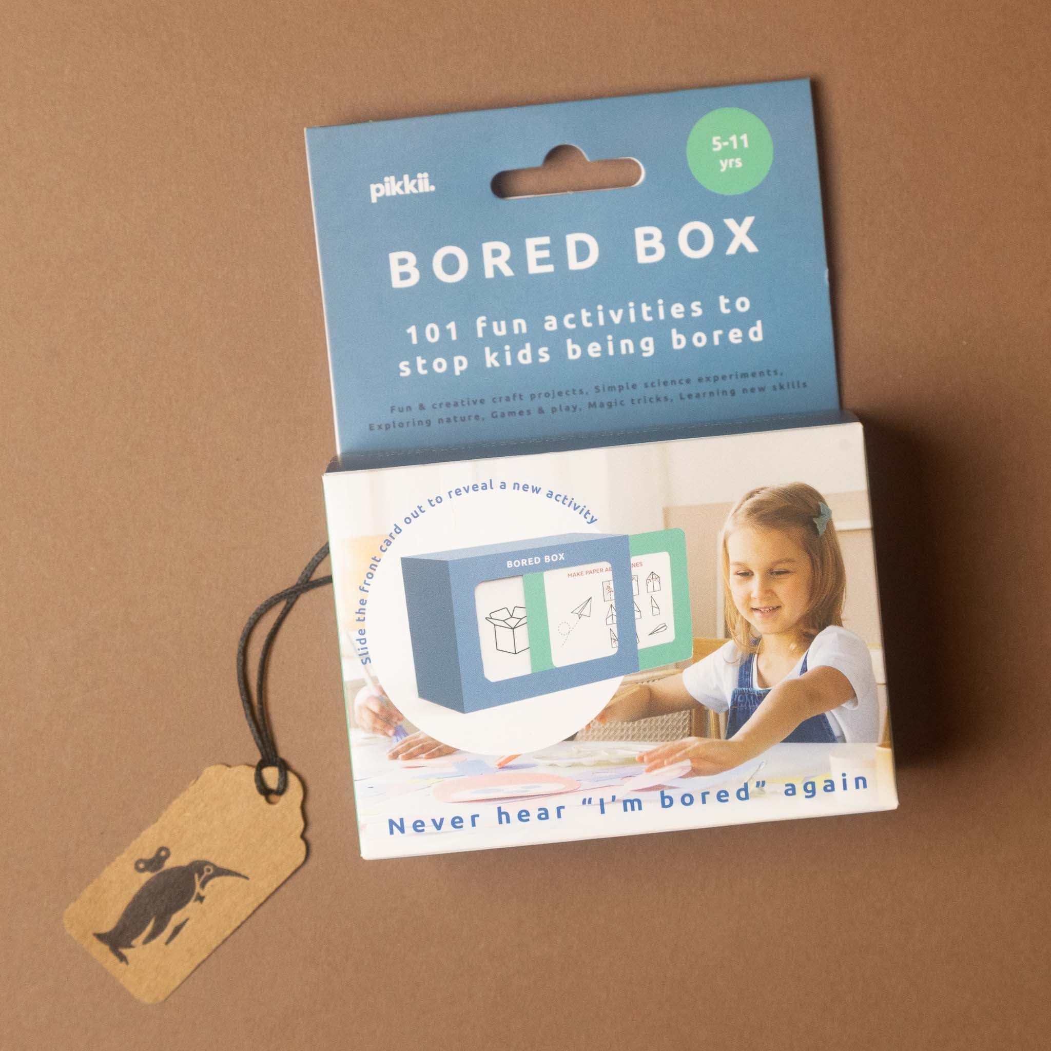 the-bored-box-kids-activity-kit on a brown background, featuring a blue header card, a printed box with a child illustration, and a small kraft paper tag with a black bird logo.