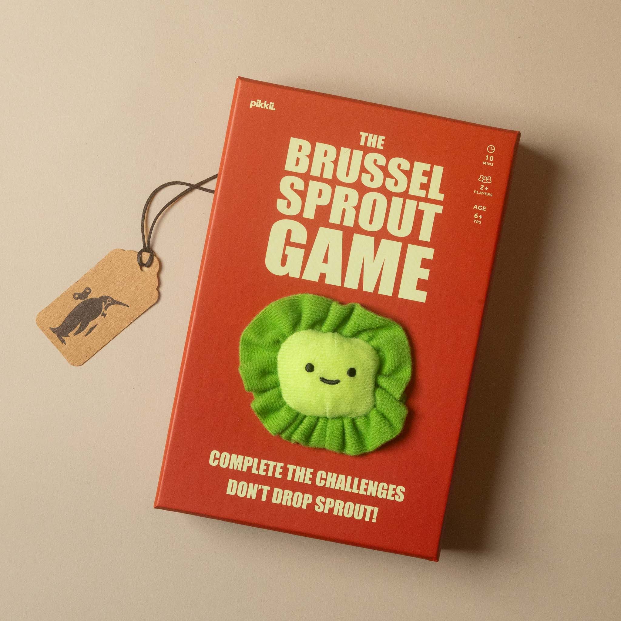 The Brussel Sprout Game box on a warm beige background, featuring a bright red cover with white text and a green plush Brussels sprout character on the front, complete with a hanging kraft paper tag.