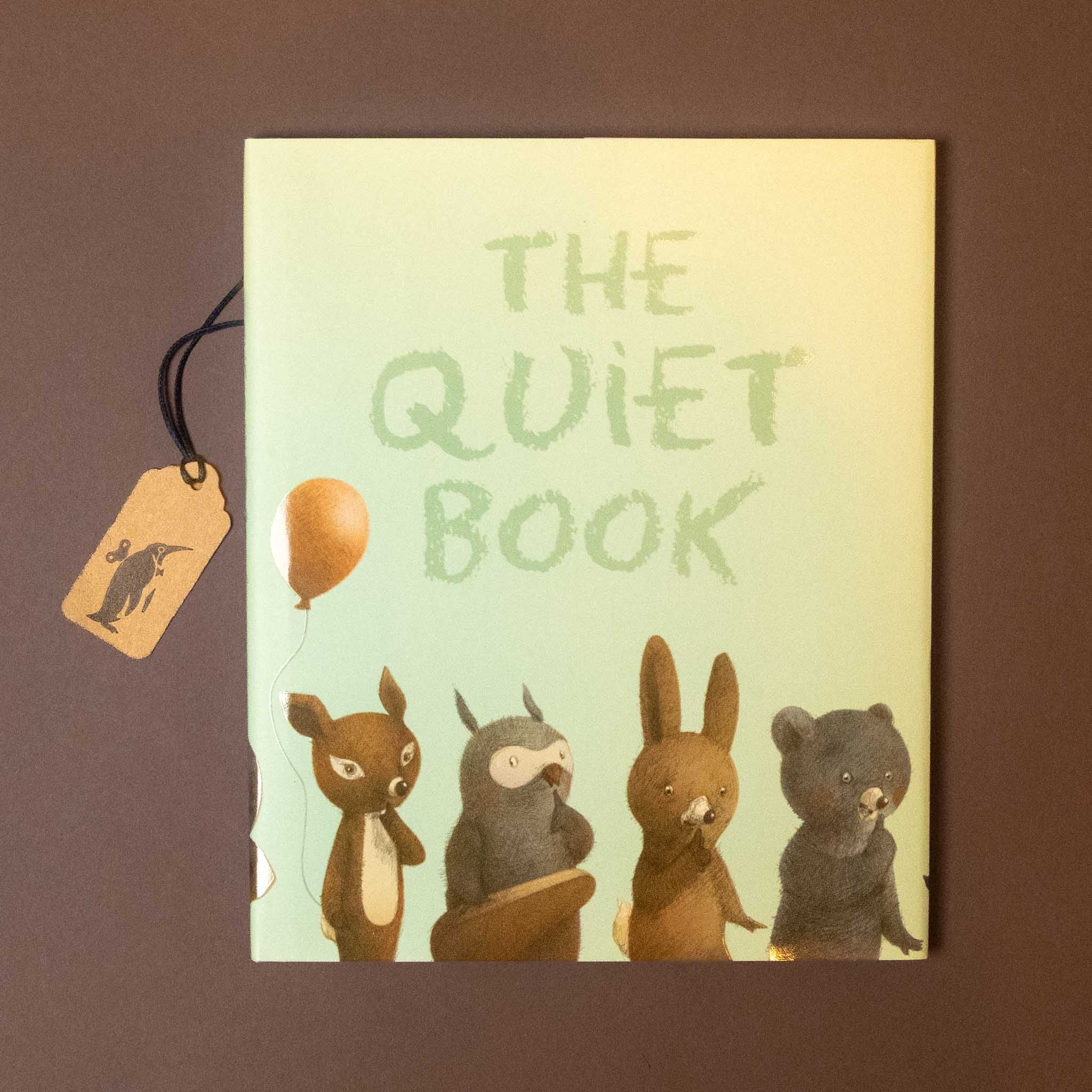 The Quiet Book with a pastel green cover featuring four cute illustrated animals and a hanging wooden tag, set against a dark brown background.