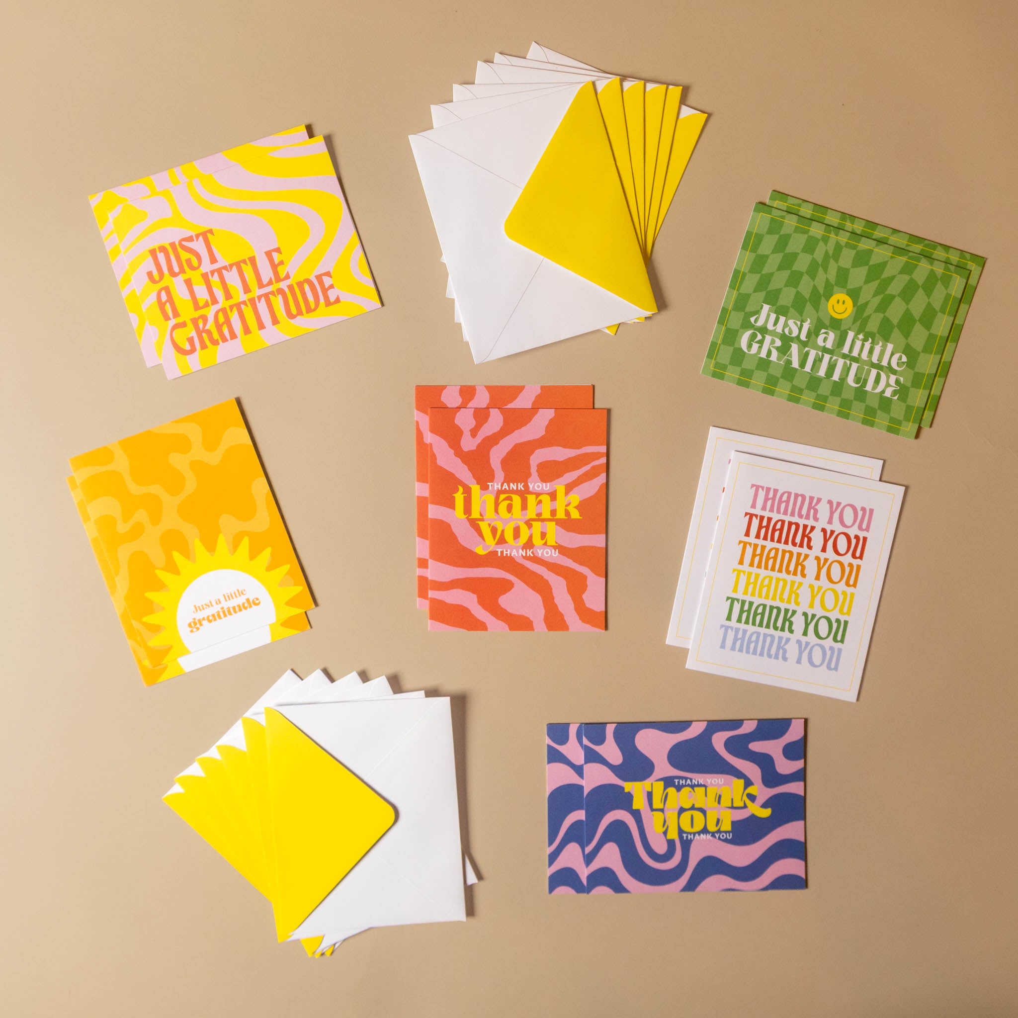 A flat-lay arrangement of multiple greeting cards and matching envelopes on a warm beige background. The cards feature colorful retro patterns including wavy lines, checkers, and stripes, with phrases like 'Just a little gratitude' and 'Thank you.