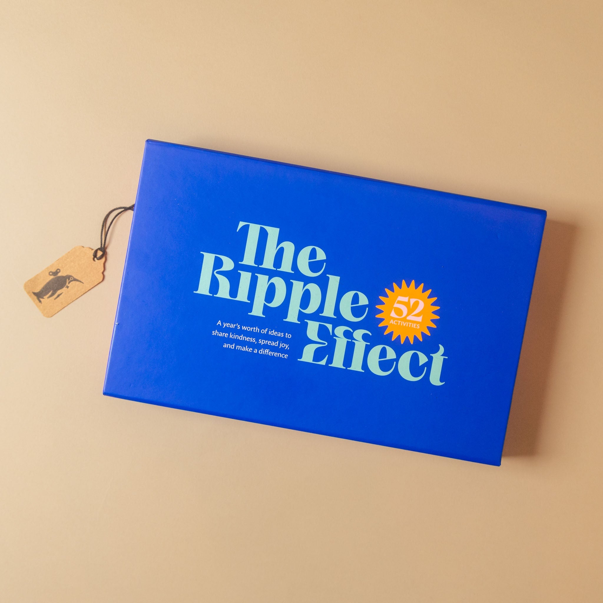 A blue rectangular product box labeled 'The Ripple Effect' with a small orange '52' burst and white text, photographed on a warm beige background. A small wooden tag hangs from the box by a dark string.