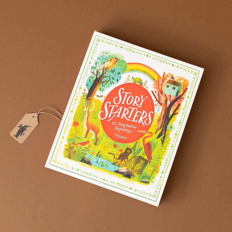 A boxed card deck titled "Story Starters" lies on a brown surface. The box has a colorful illustrated cover with a rainbow, trees, animals, and a house, plus a small hanging tag attached by a string.