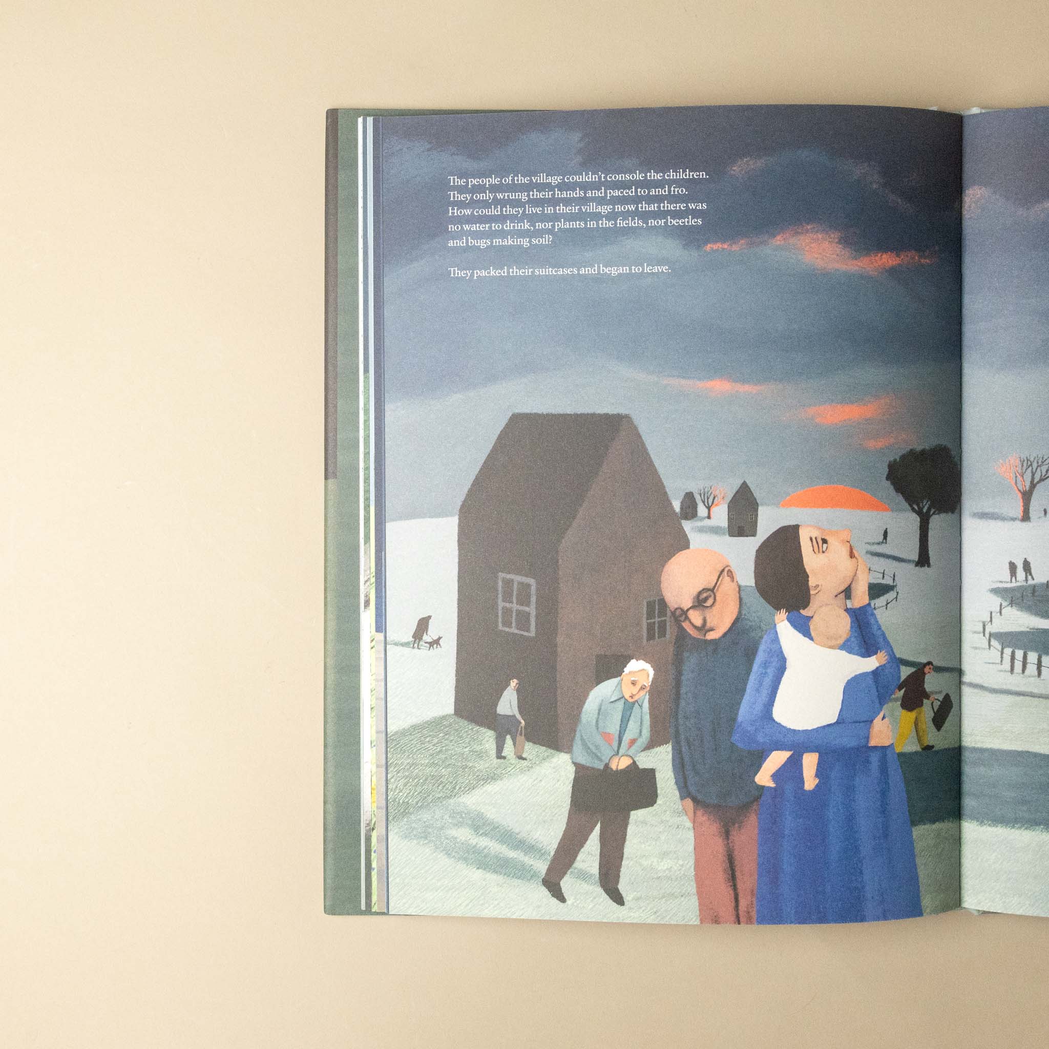 An open children's book lying on a beige surface, showing a painted winter village scene with a house, people, and a dark blue sky. The left page includes a block of text at the top.