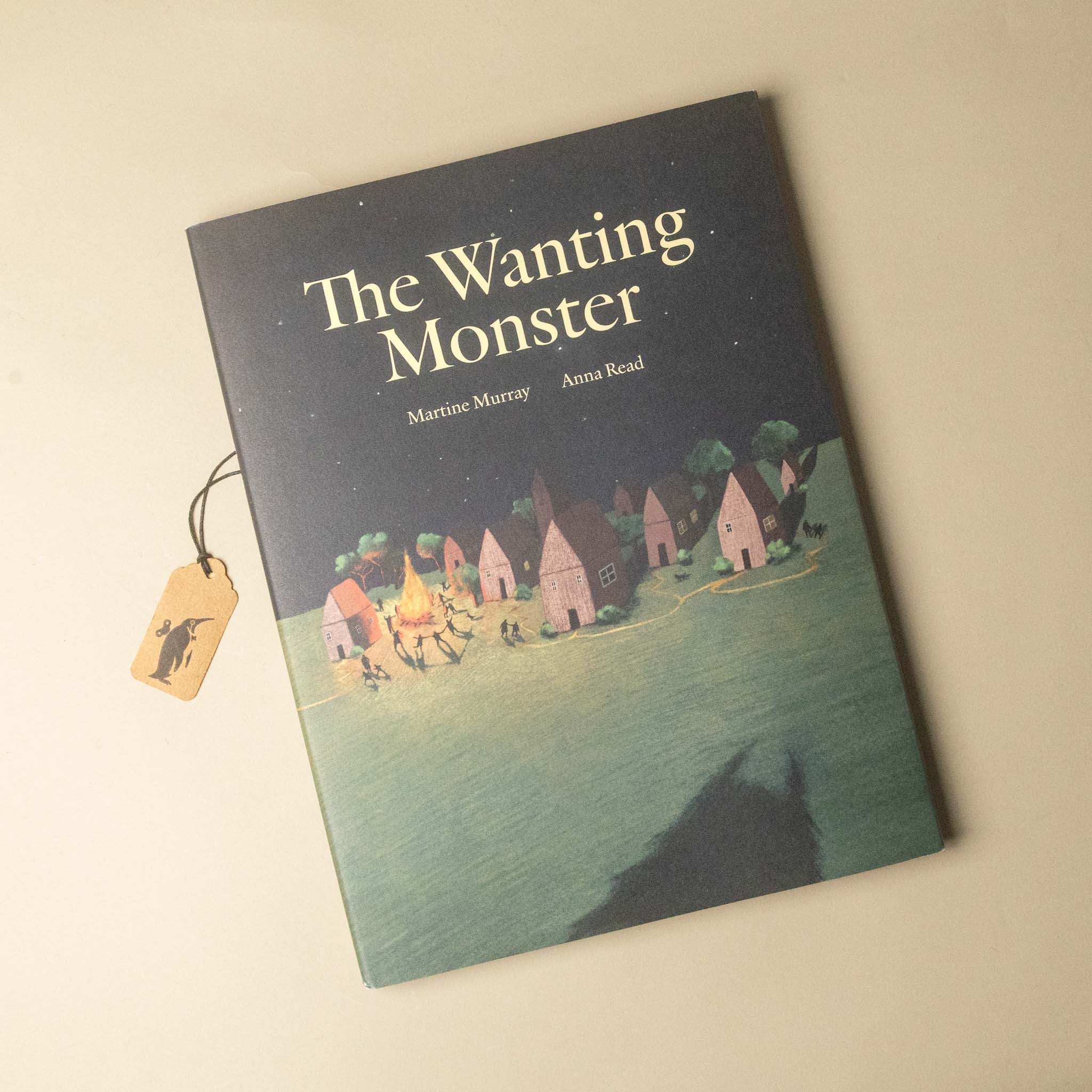 The Wanting Monster book by Martine Marczak and Anna Read, featuring a dark illustrated cover with a small village and a wolf silhouette, shown on a light beige background with a small wooden tag attached.