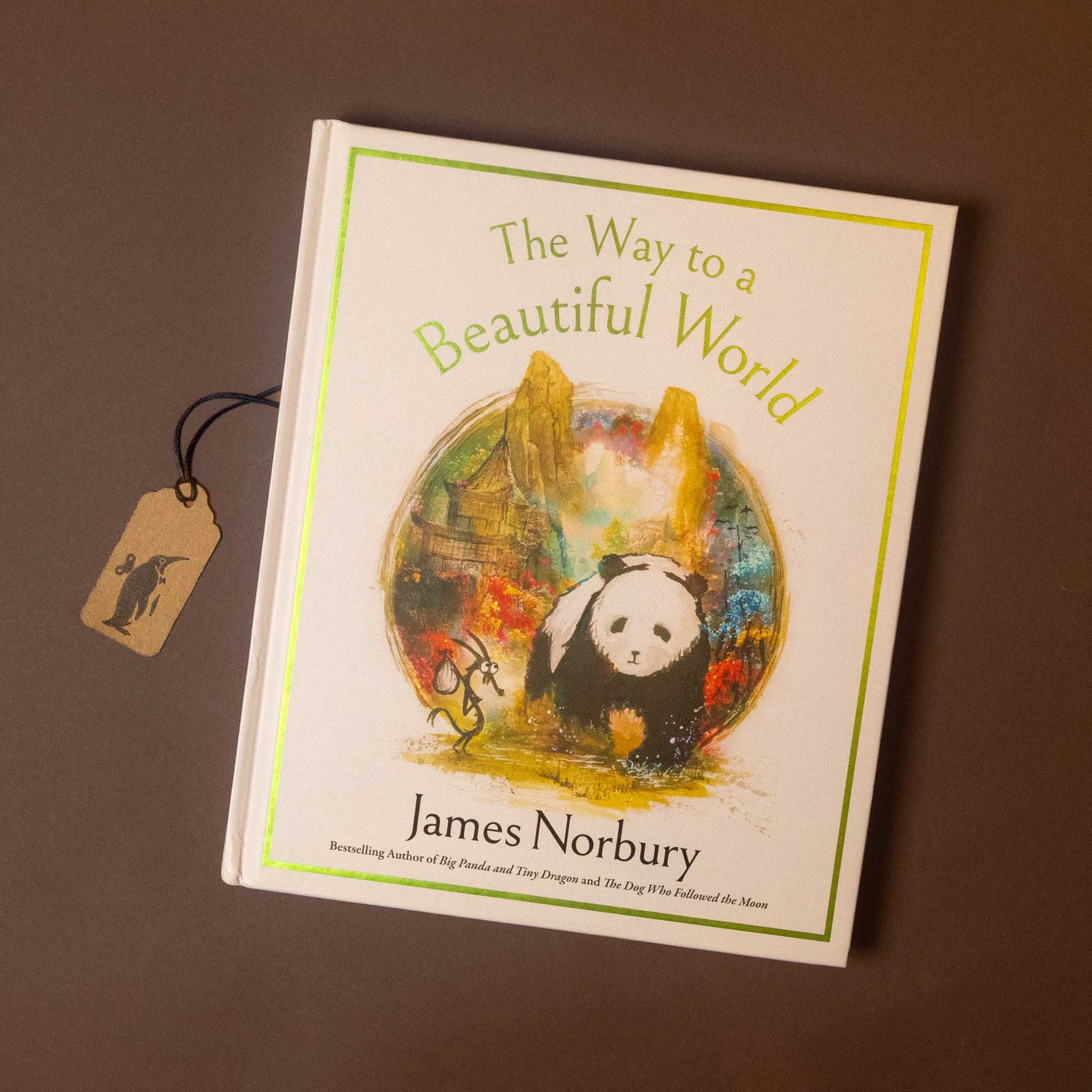 The Way to a Beautiful World book by James Norbury, featuring a panda illustration on the cover, displayed on a dark brown background with a small paper tag attached.