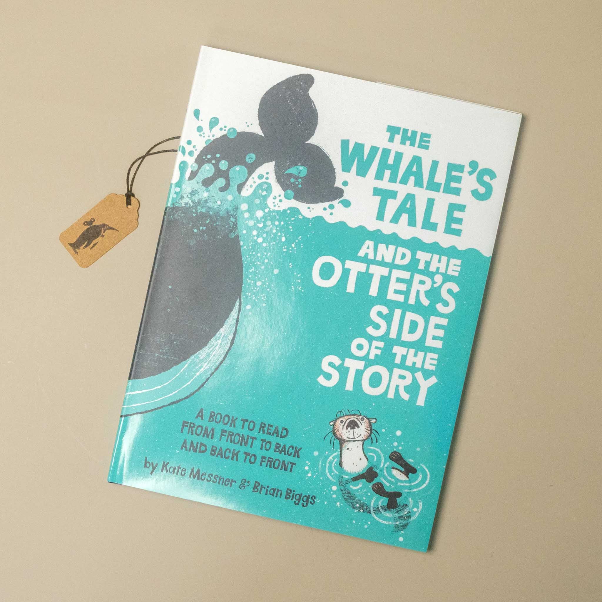 A children's book titled "The Whale's Tale and the Otter's Side of the Story" lies on a beige surface, angled slightly to the right. The cover is white and turquoise with a whale tail and an otter illustration.