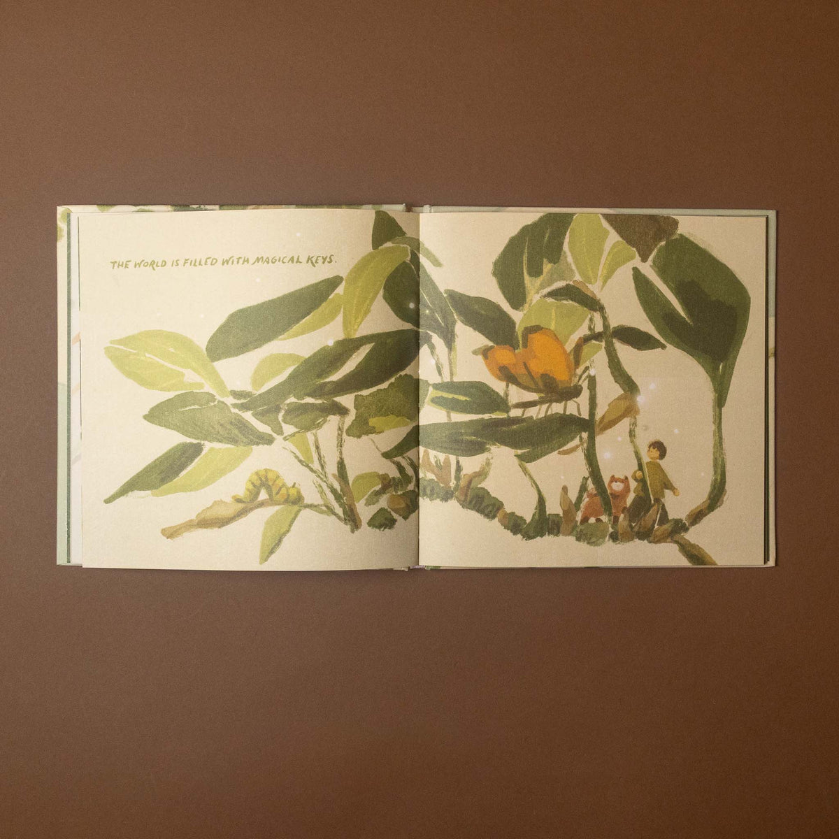 An open illustrated book spread showing botanical artwork with green leaves, a yellow-orange flower, and small figures, photographed against a plain brown background.