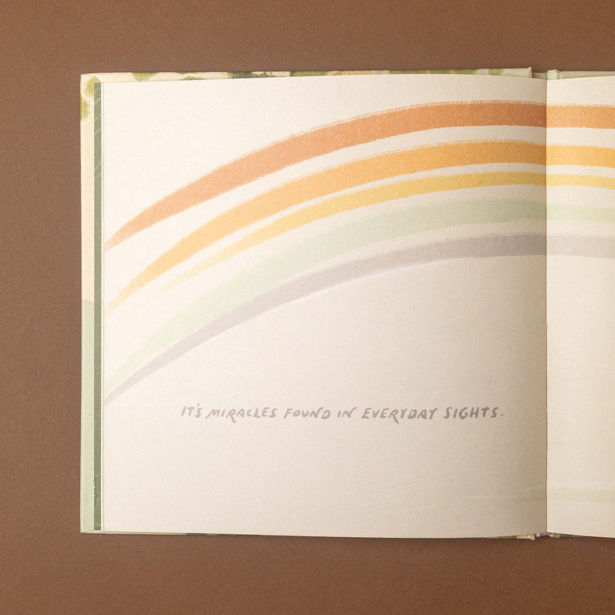 An open book or booklet spread on a brown background, featuring a pastel rainbow stripe illustration across the pages and the handwritten text, 'IT'S MIRACLES FOUND IN EVERYDAY SIGHTS.'.