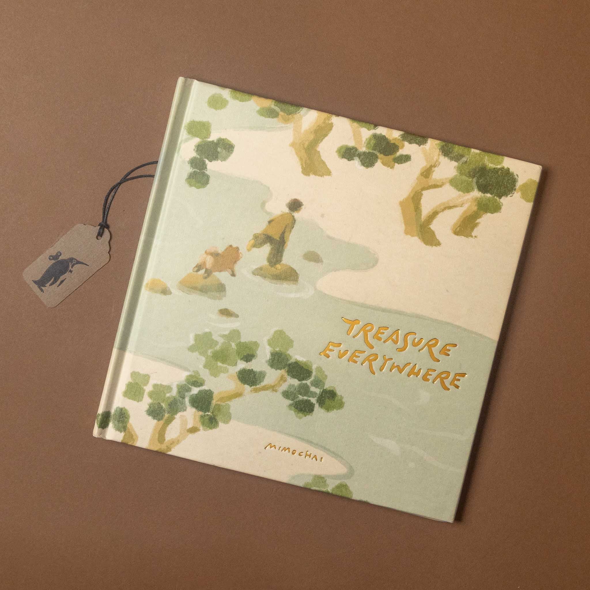 A hardcover art book titled 'Treasure Everywhere' by Munchai, featuring a whimsical illustration of a figure and a dog on a light green landscape with gold lettering, displayed on a brown background with a small branded hangtag.