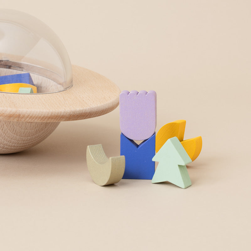 A set of pastel-colored wooden geometric blocks arranged on a beige surface, with a large wooden plate and clear dome container partially visible in the background.