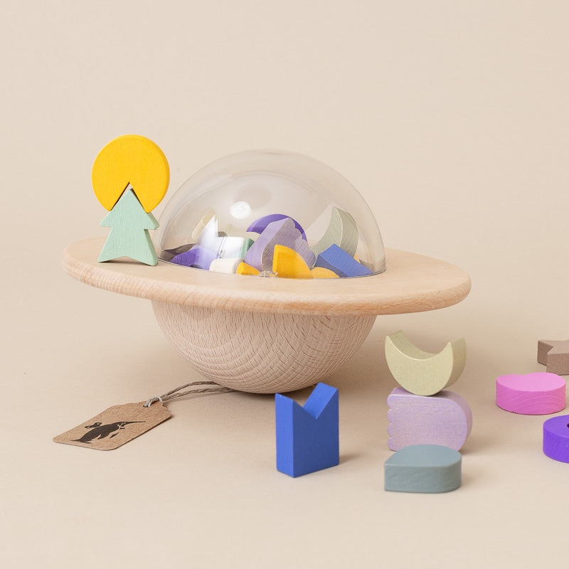 A wooden planet-shaped toy bowl with a clear dome lid, filled with pastel geometric shapes, on a warm beige background with additional loose wooden blocks and a hanging tag nearby.