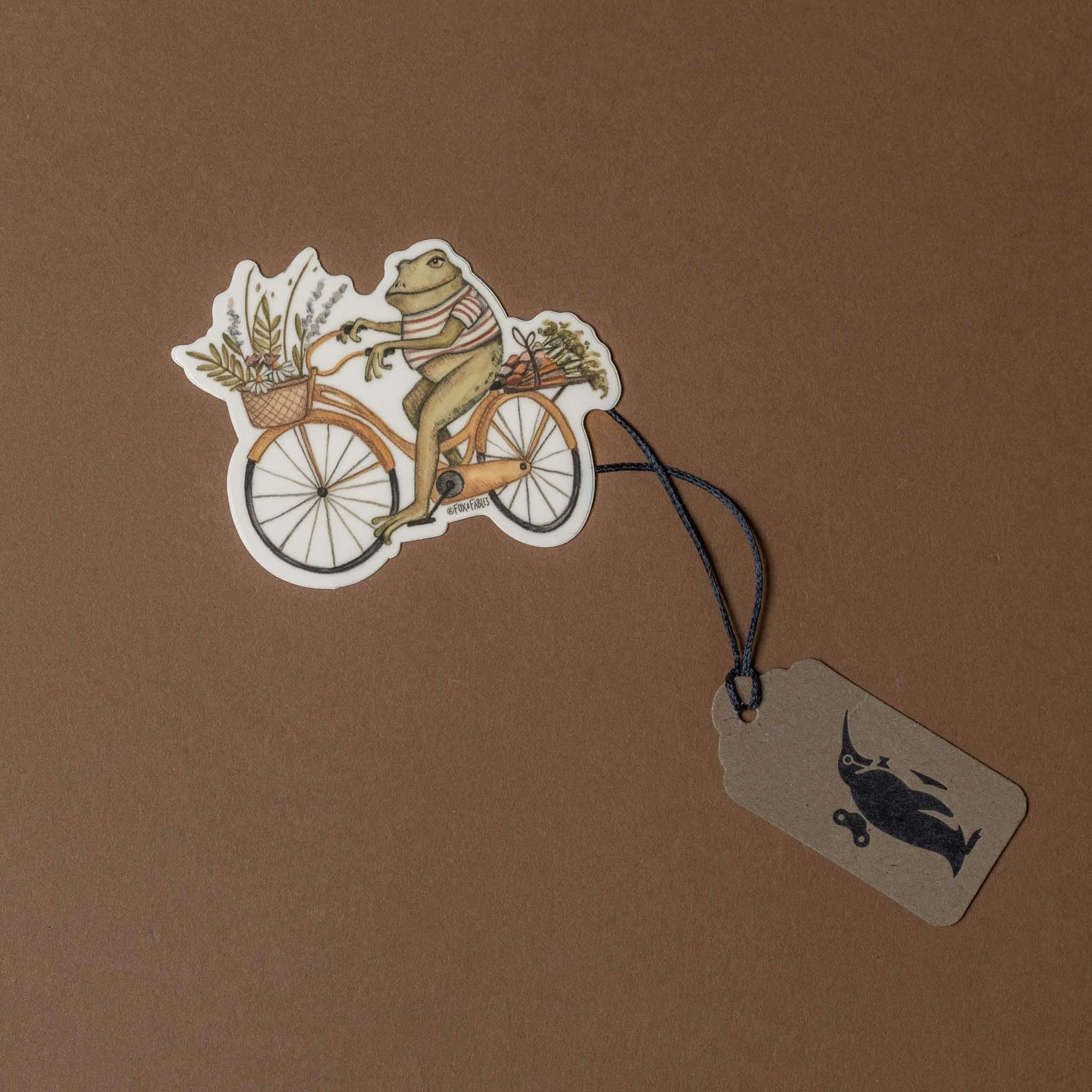 Frog on a bicycle with a basket, attached to a tag on a brown background