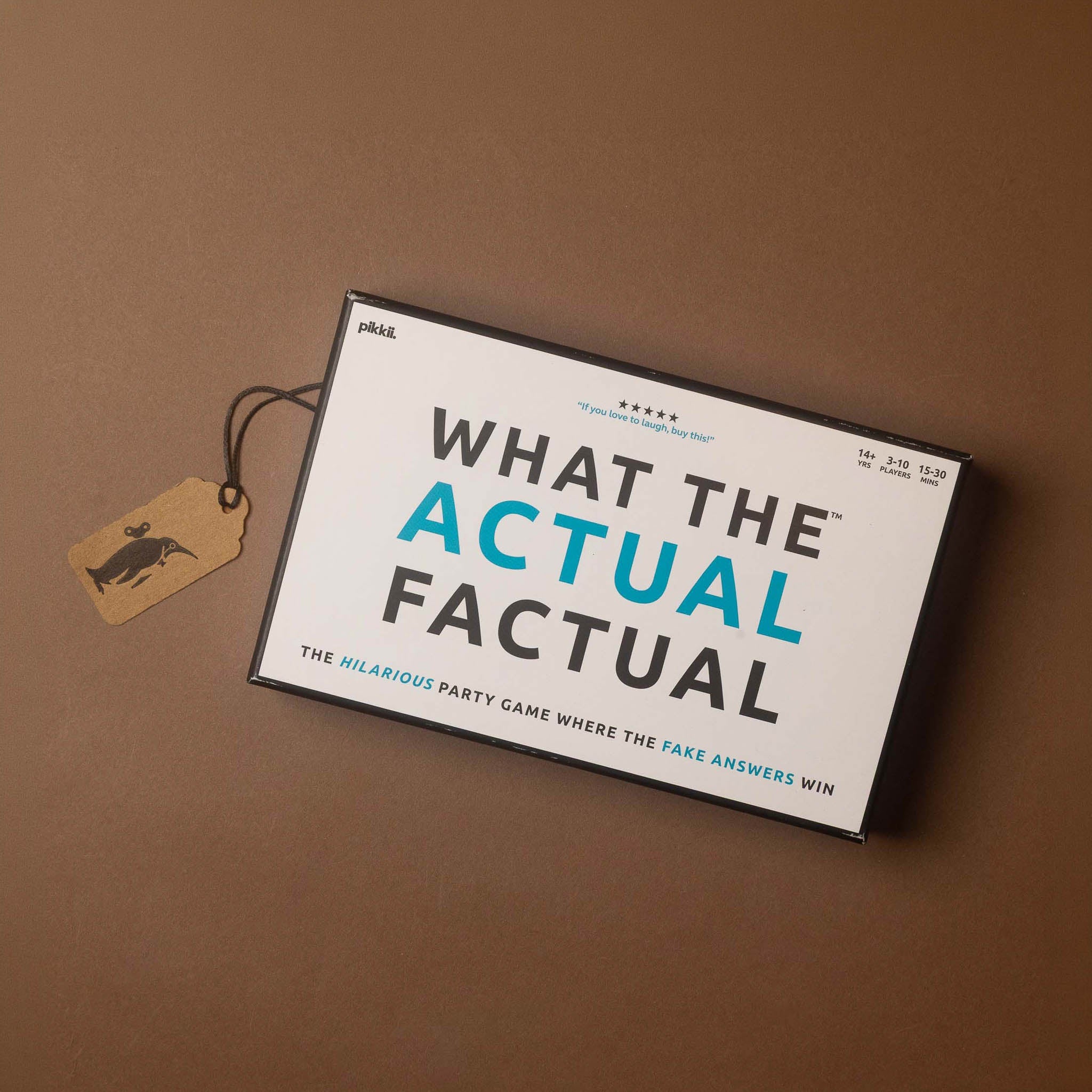 The product box for the party game 'What the Actual Factual' is shown at an angle on a solid brown background, complete with a small paper tag attached.