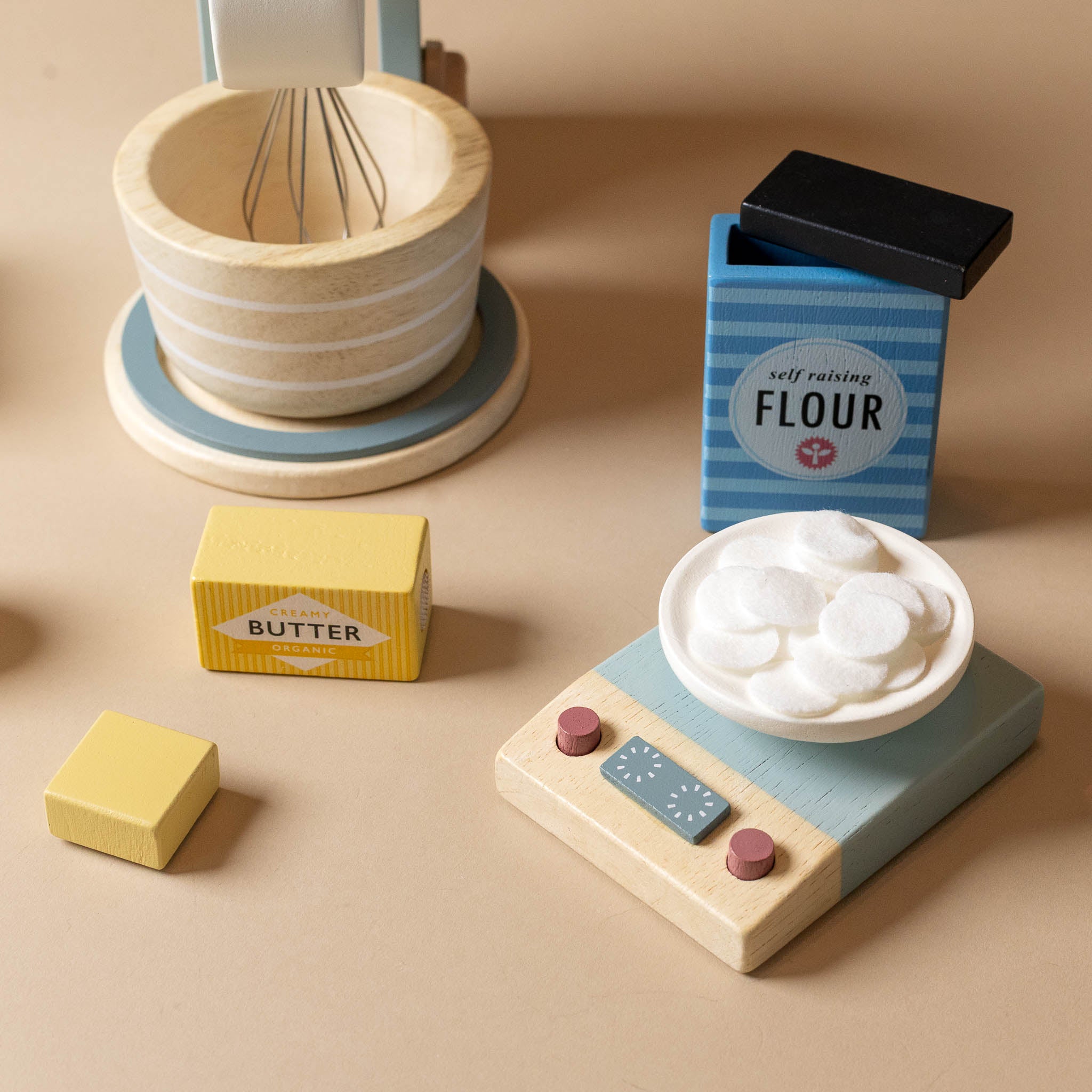 wooden-bake-a-cake-play-set Wooden play kitchen set with flour, butter, and a whisk on a beige background