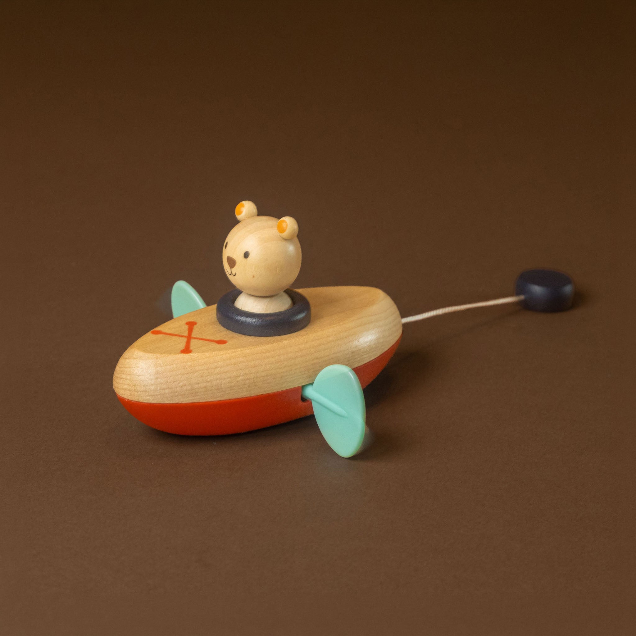 wooden-clockwork-float-boat-bear on top, featuring a light natural wood body, orange-red bottom, mint green fins, and a dark blue tail, set against a solid brown background.