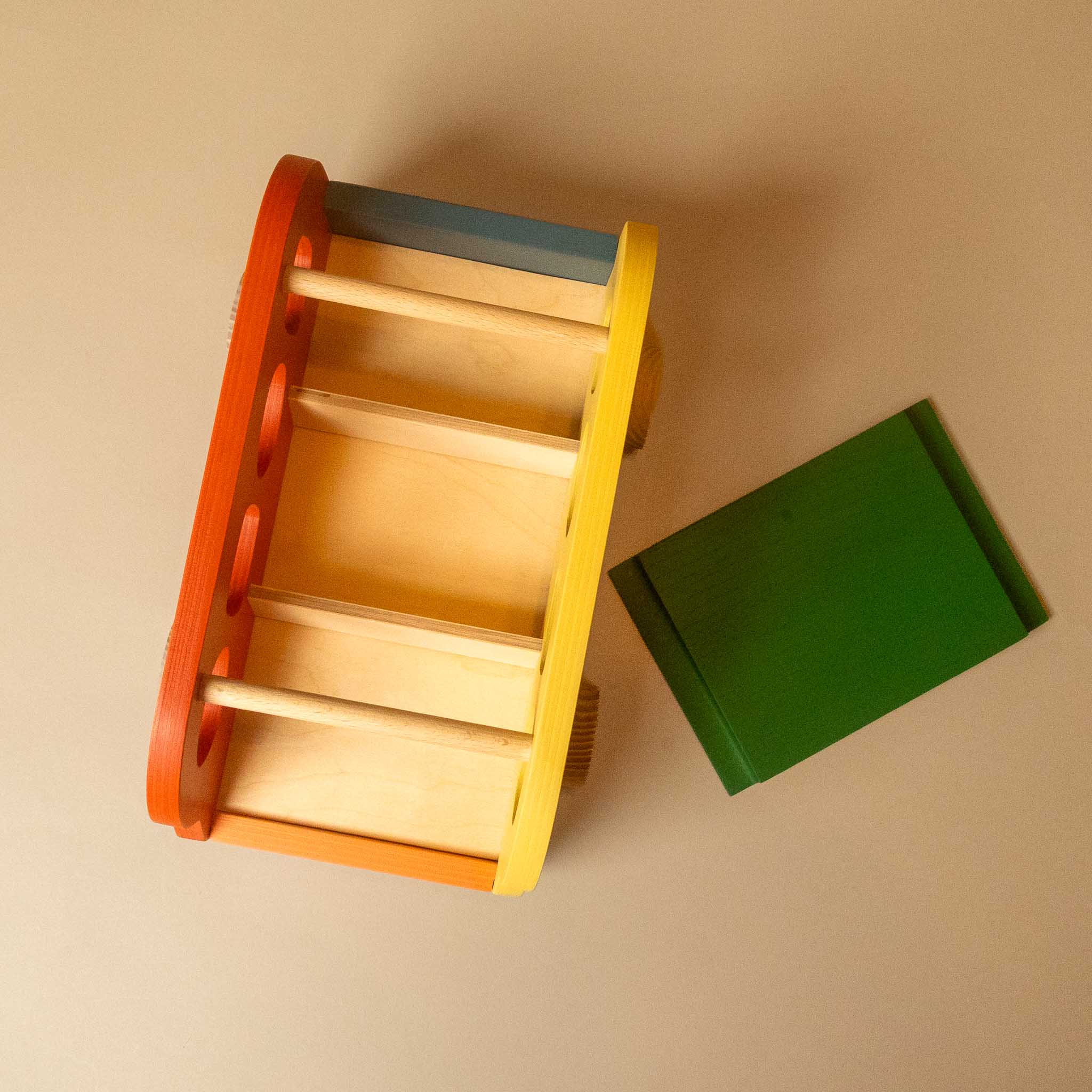 A wooden toy box with colorful painted edges in orange, yellow, blue, and brown, shown alongside a green rectangular wooden block on a plain beige background.