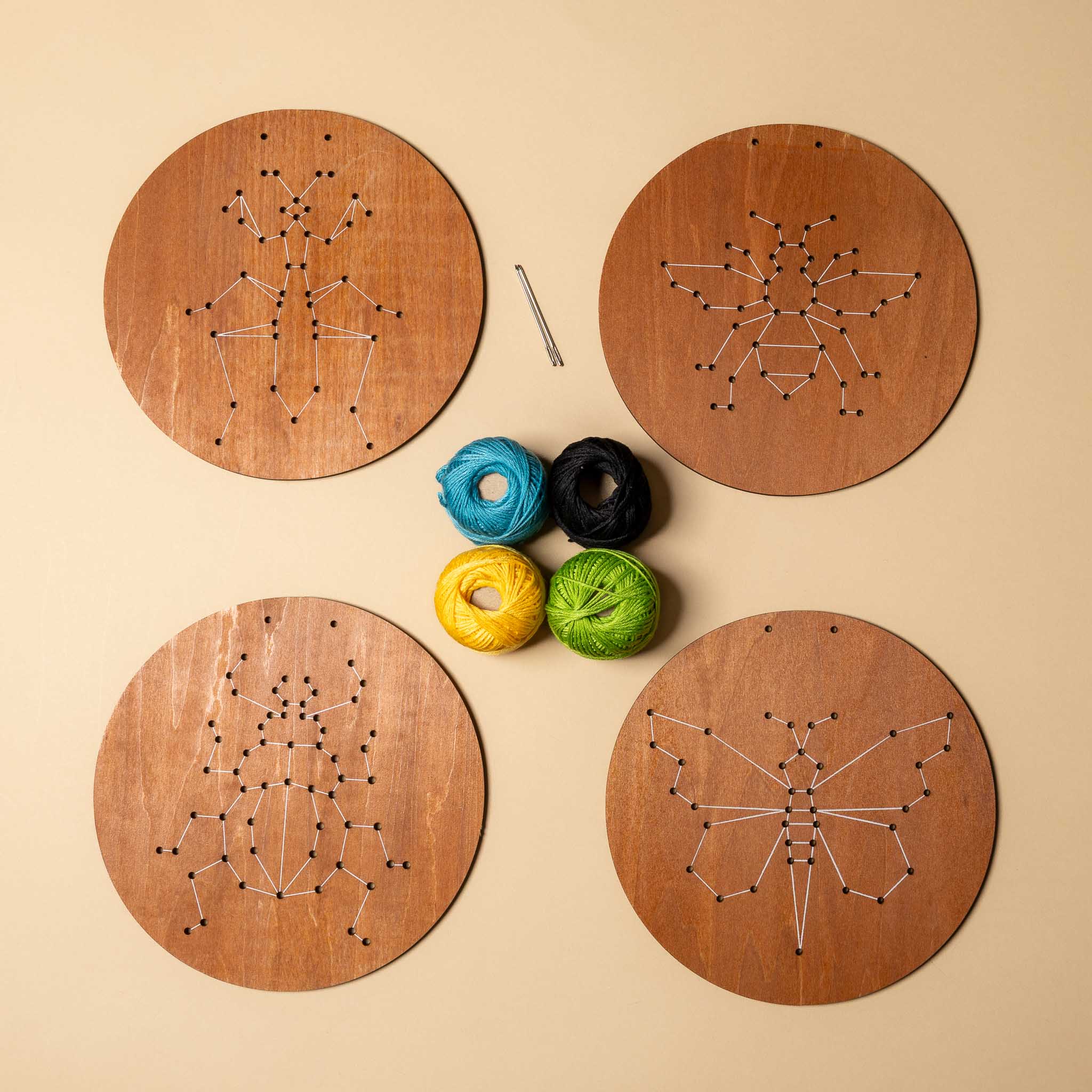 wooden-embroidery-kit-insects Four wooden boards with string art patterns and colorful yarn on a beige background