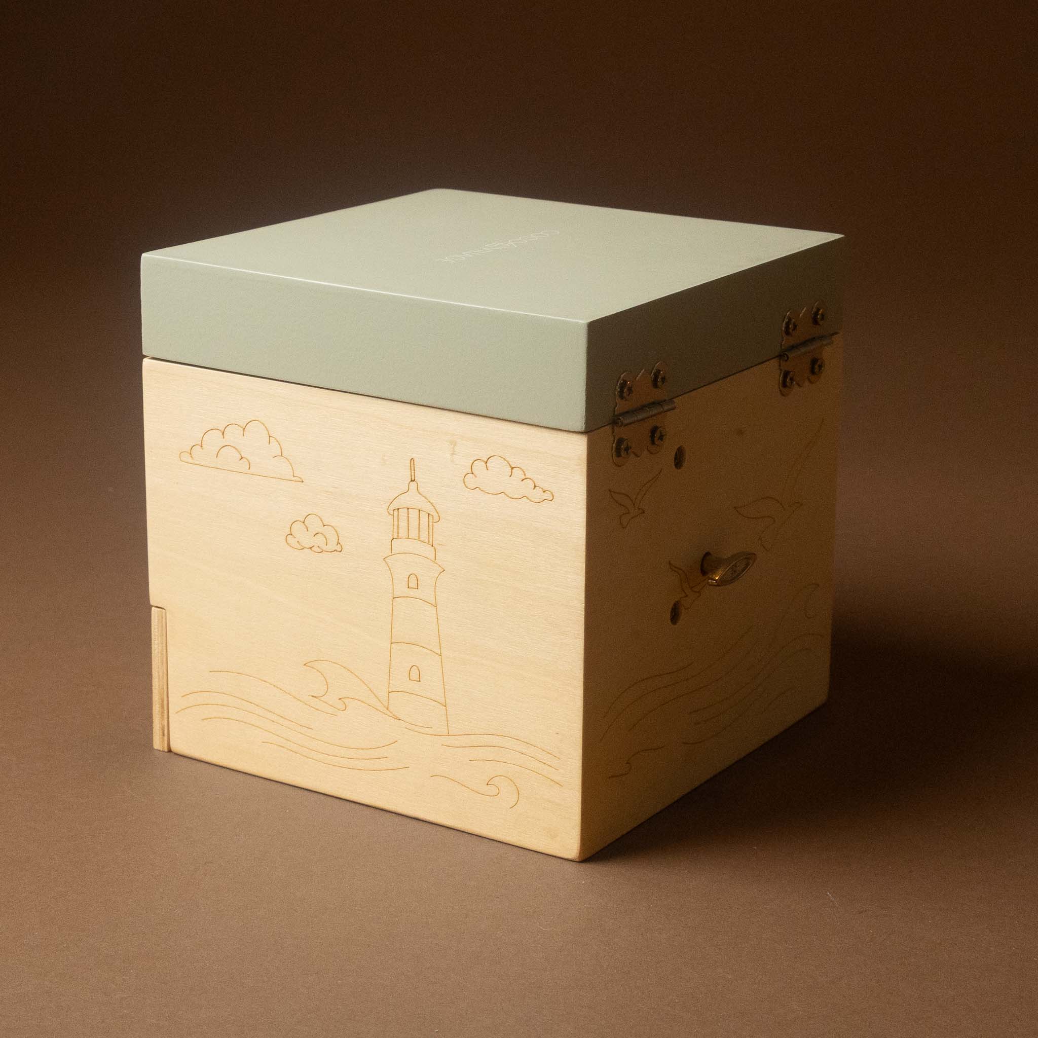 wooden-musical-treasures-box-by-the-sea with a light natural wood front panel engraved with a lighthouse, clouds, and waves, and a pale sage green lid. The box has small metal hinges on the lid and a small metal handle on the side, photographed on a solid brown background.
