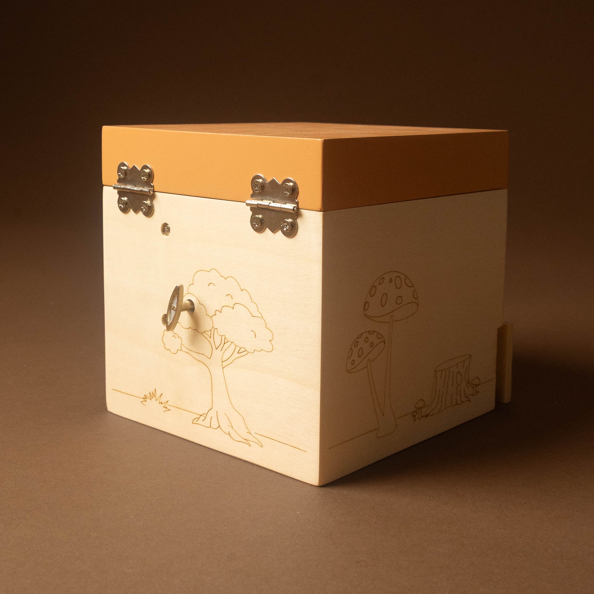 wooden-musical-treasures-box-nature with a light natural wood body, a warm orange-brown lid, and engraved forest illustrations including a tree and mushrooms. It has metal corner hinges and a small keyhole cutout on the front, photographed on a plain brown studio background.