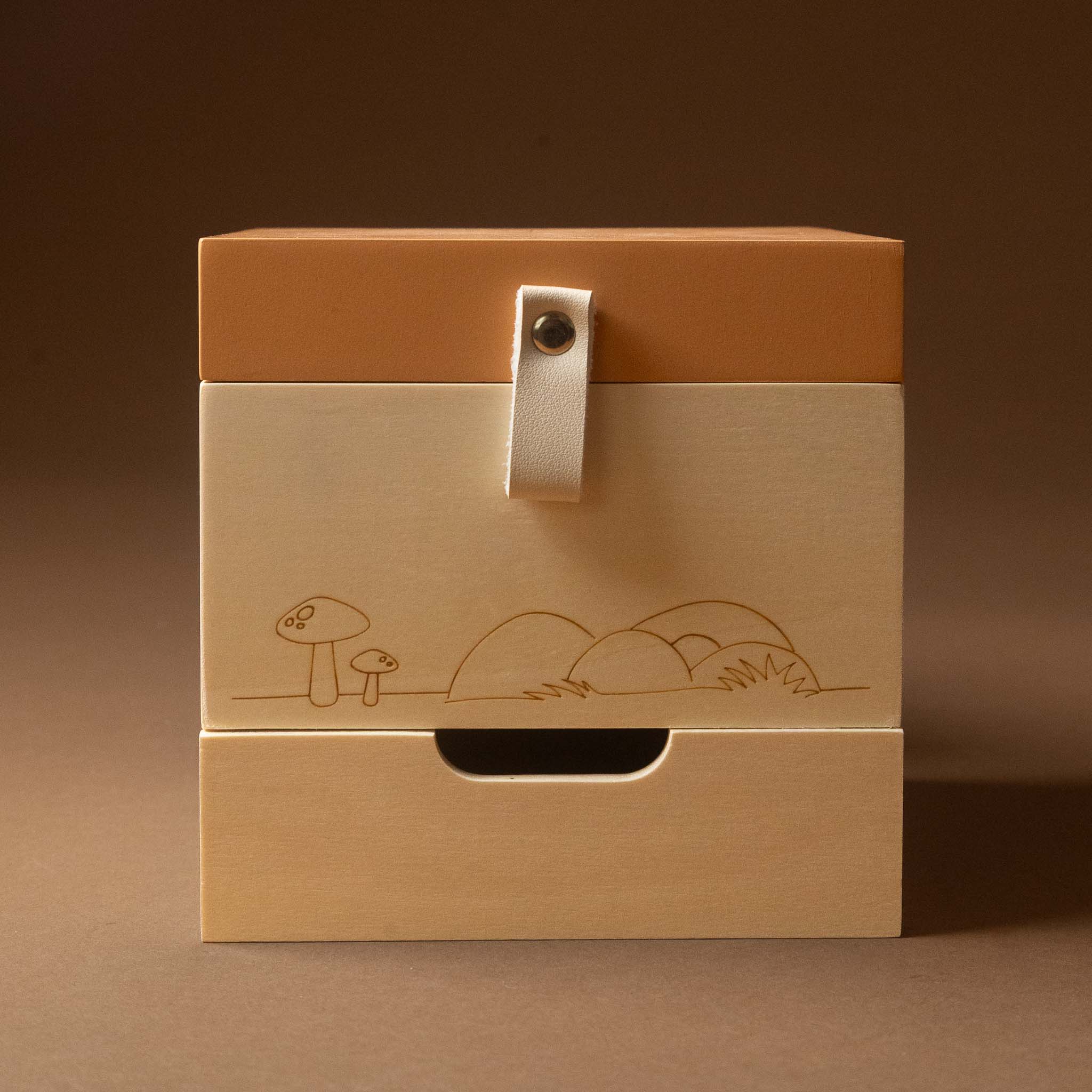 wooden-musical-treasures-box-nature with a warm brown lid, brushed beige front panel, and a small white strap closure. The front is engraved with a simple mushroom and hill illustration, set against a solid brown background.
