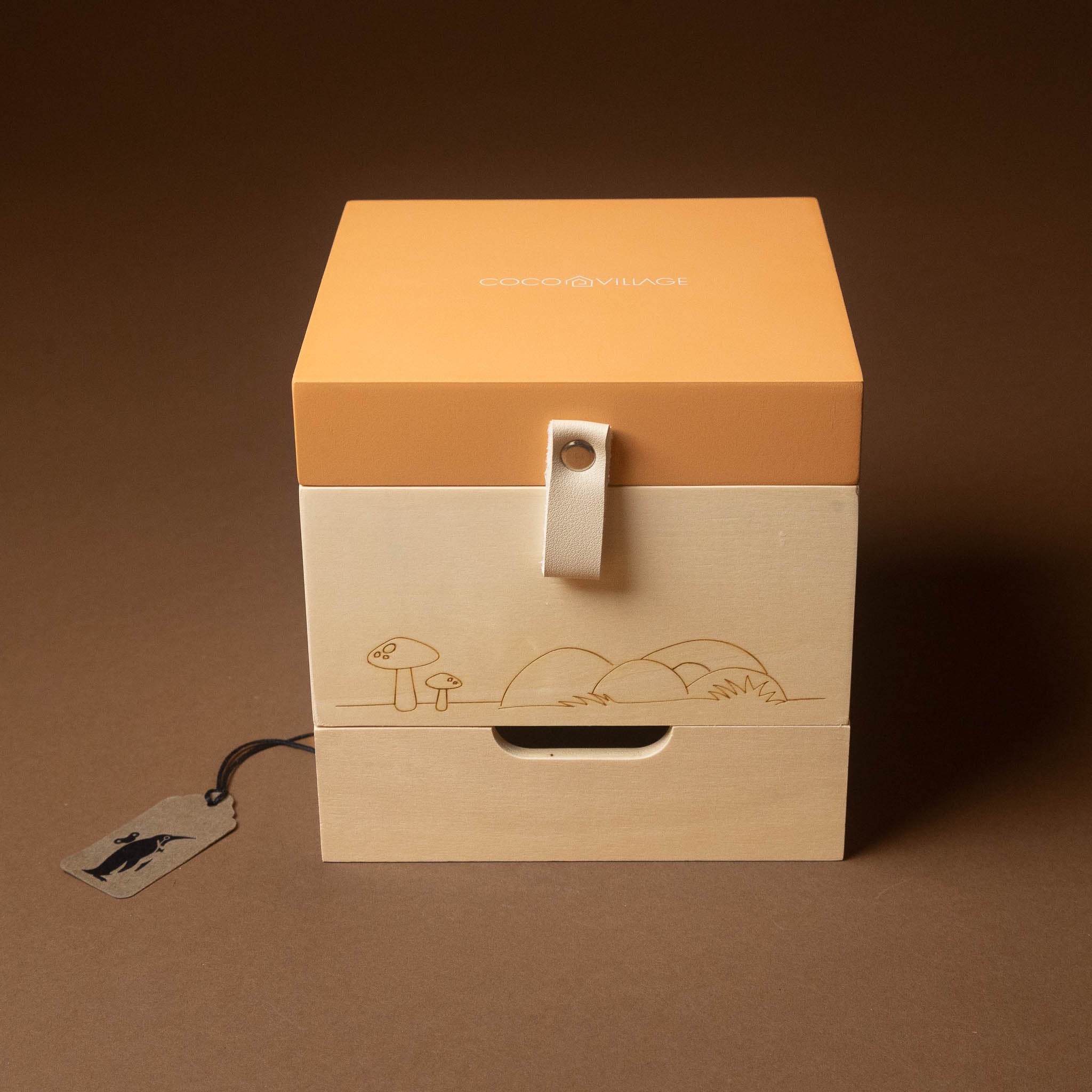 wooden-musical-treasures-box-nature with a warm orange lid and natural light wood drawers, featuring a white hanging loop and simple line-art illustrations on the front. A small branded tag hangs from a black cord on the left against a solid brown background.