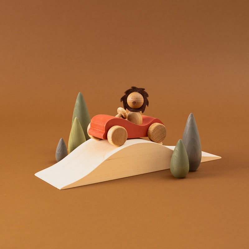 A wooden toy scene showing a small lion figure driving a red car on a curved white ramp, surrounded by simple pine trees, against a warm brown studio background.