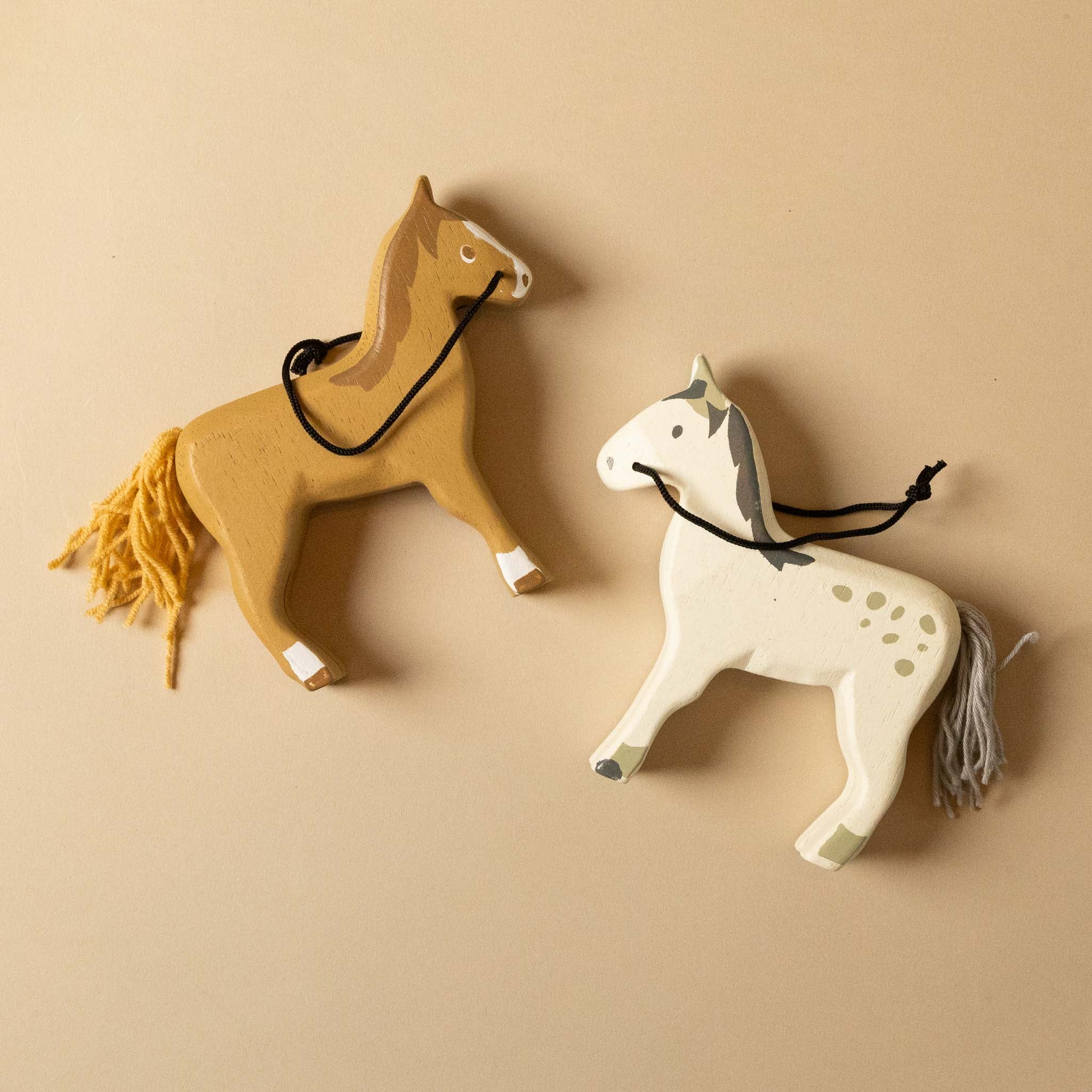 wooden-stable-play-set Two toy horses, one brown and one white with gray spots, on a beige background.