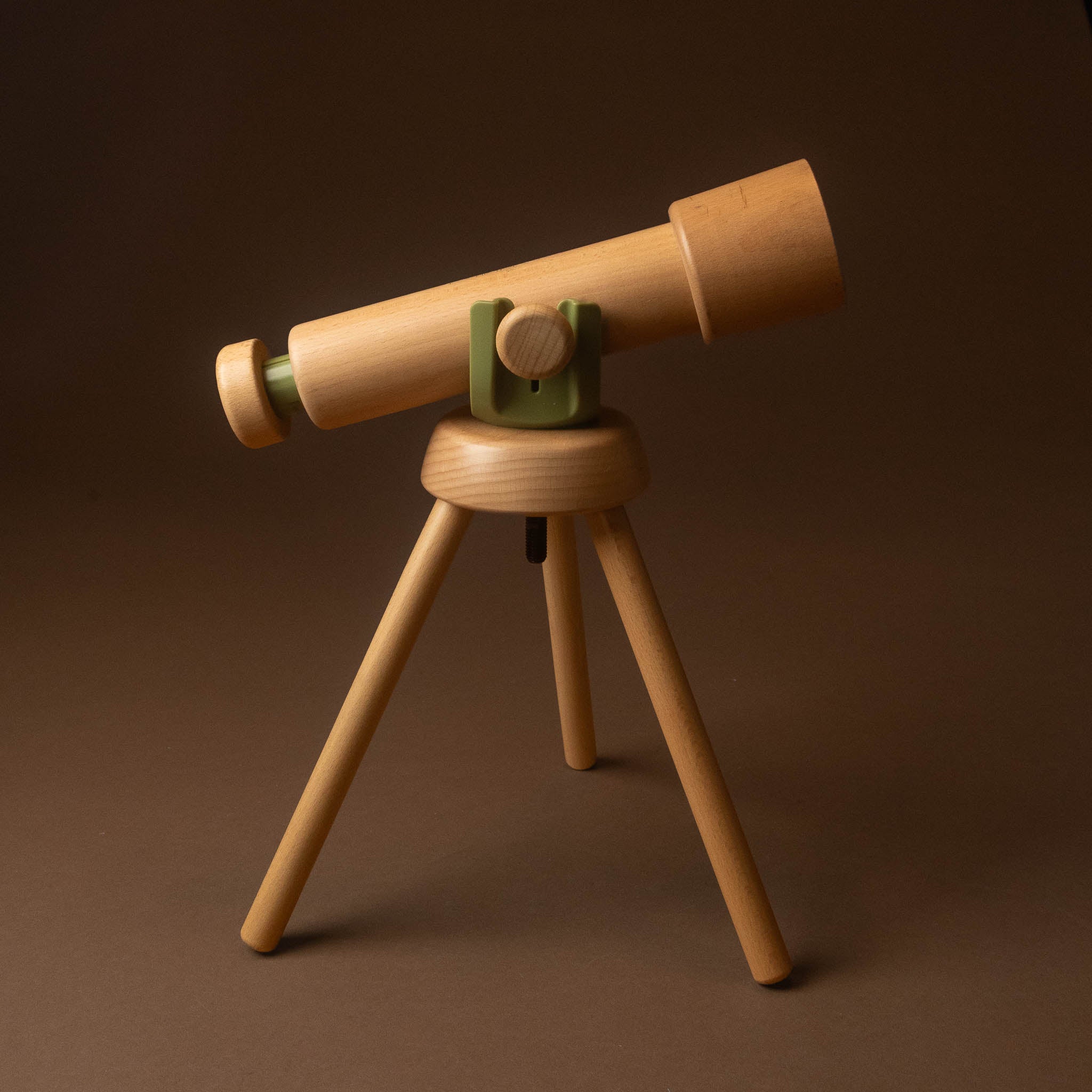 A wooden toy or model telescope mounted on a three-legged tripod stand, photographed against a dark brown studio background.