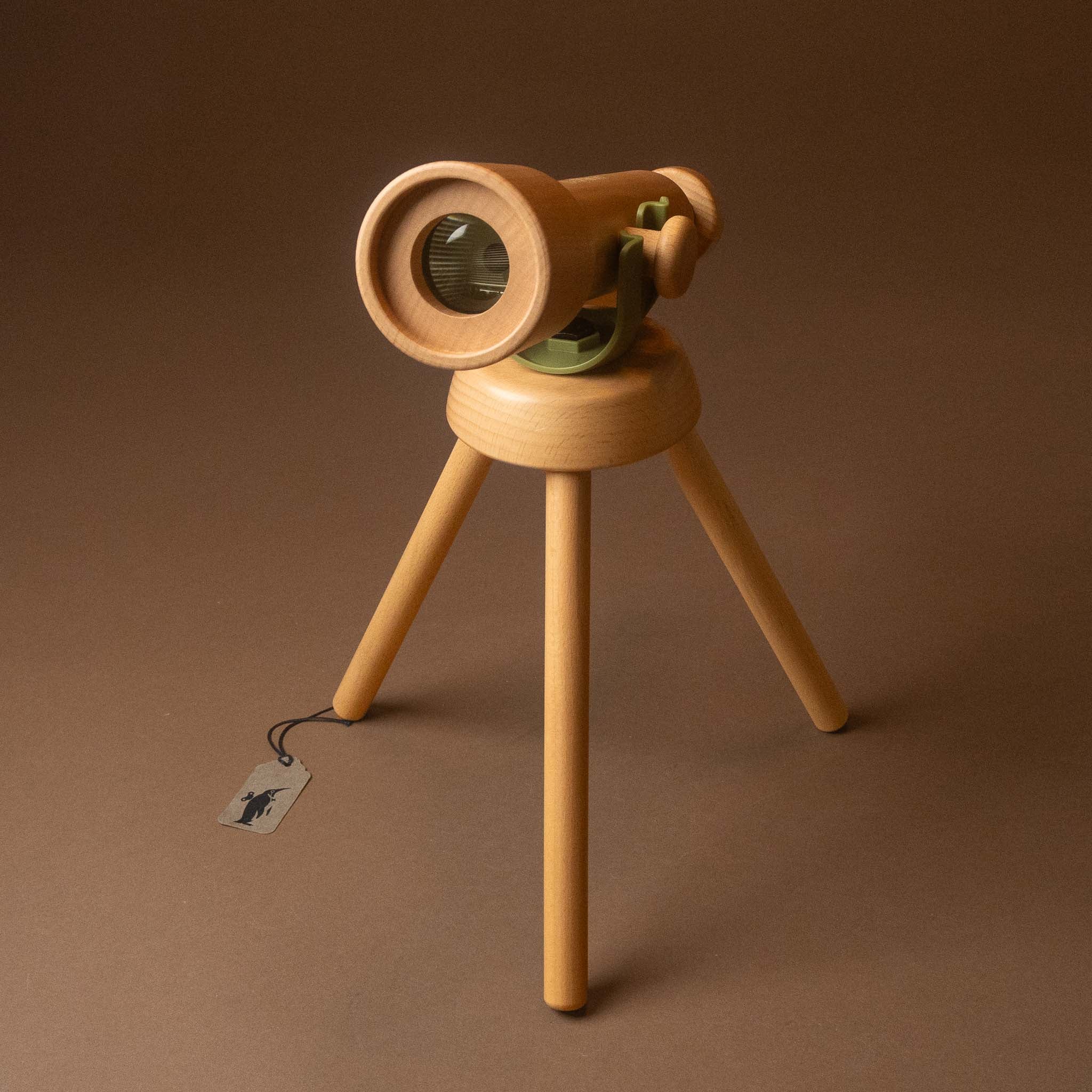 A wooden toy telescope mounted on a three-legged tripod stand, photographed against a solid brown studio background. A small paper tag hangs from the tripod leg.